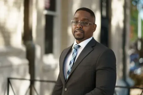 Professional in a dark suit and striped tie standing in a bright outdoor corridor.