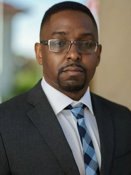 Portrait of a man in glasses wearing a dark suit and blue patterned tie, standing indoors.