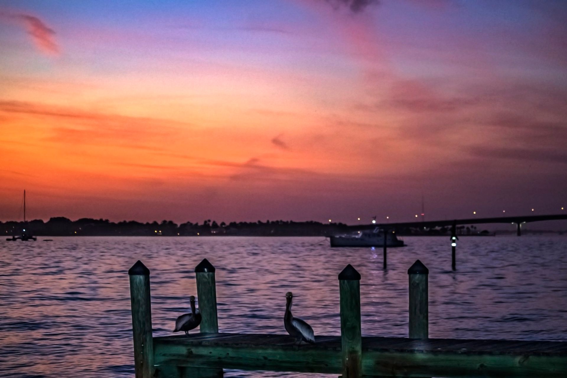 Two pelicans perch on wooden pilings overlooking a harbor at sunset with a vibrant orange and purple sky.