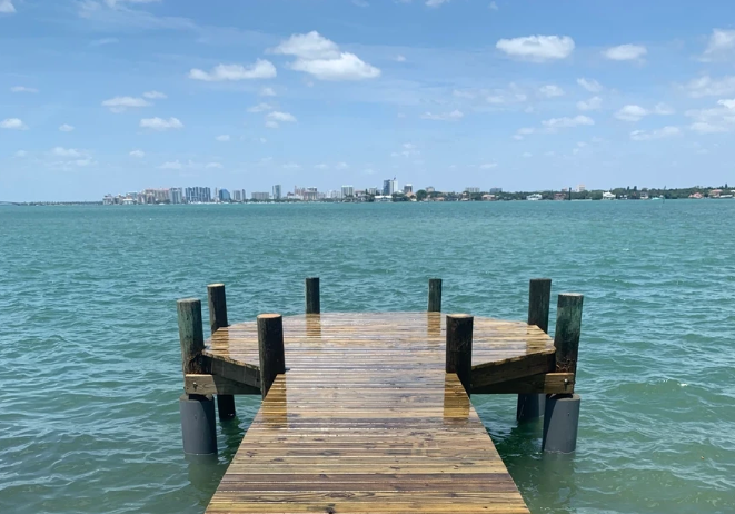 A wooden pier extends into calm, turquoise water under a blue, cloudy sky, with a distant city skyline on the horizon.