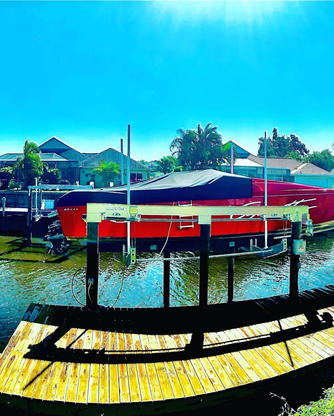A red boat with a black cover sits on a boat lift in a canal near houses under a bright, sunny sky.