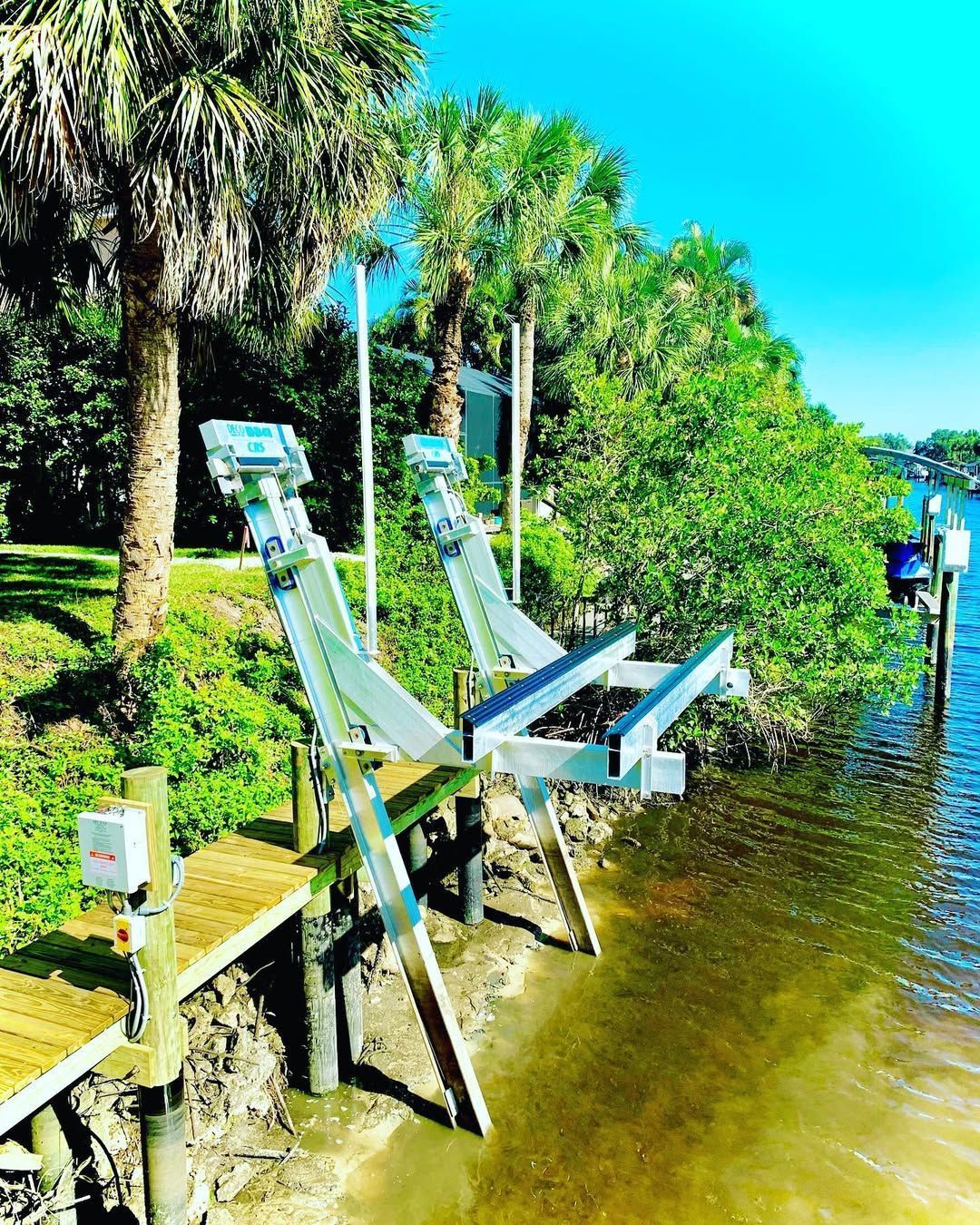 A dual boat lift mechanism stands over the water's edge, mounted on wooden pilings near palm trees and green vegetation.