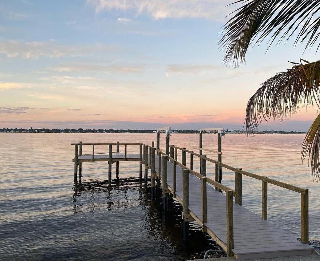 A wooden pier stretches into calm, wide water under a soft, pastel-colored sunset sky, with palm fronds in the foreground.