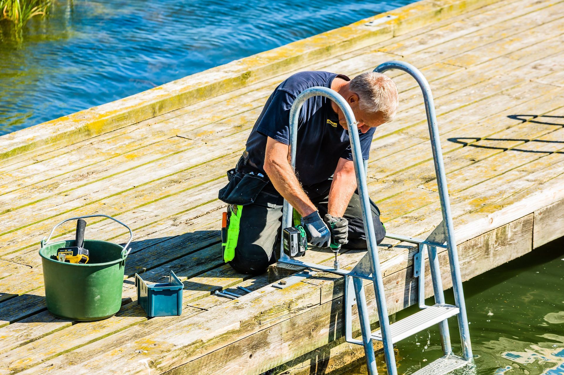 A person in work clothes kneels on a wooden dock, using a drill to install a metal swimming ladder by the water.