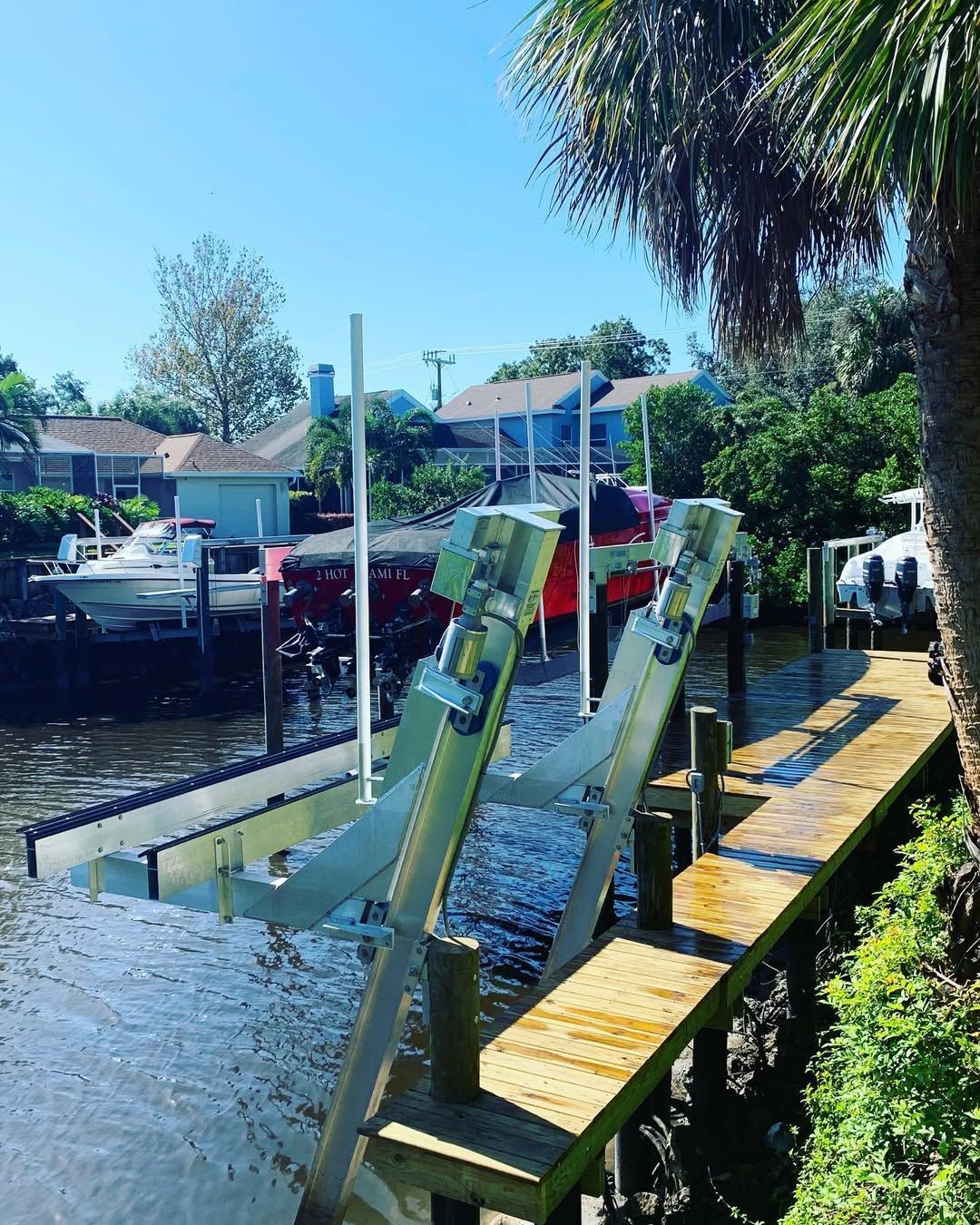 Floating dock by calm canal with green and yellow kayak launch rails, palm trees, and houses in the background.