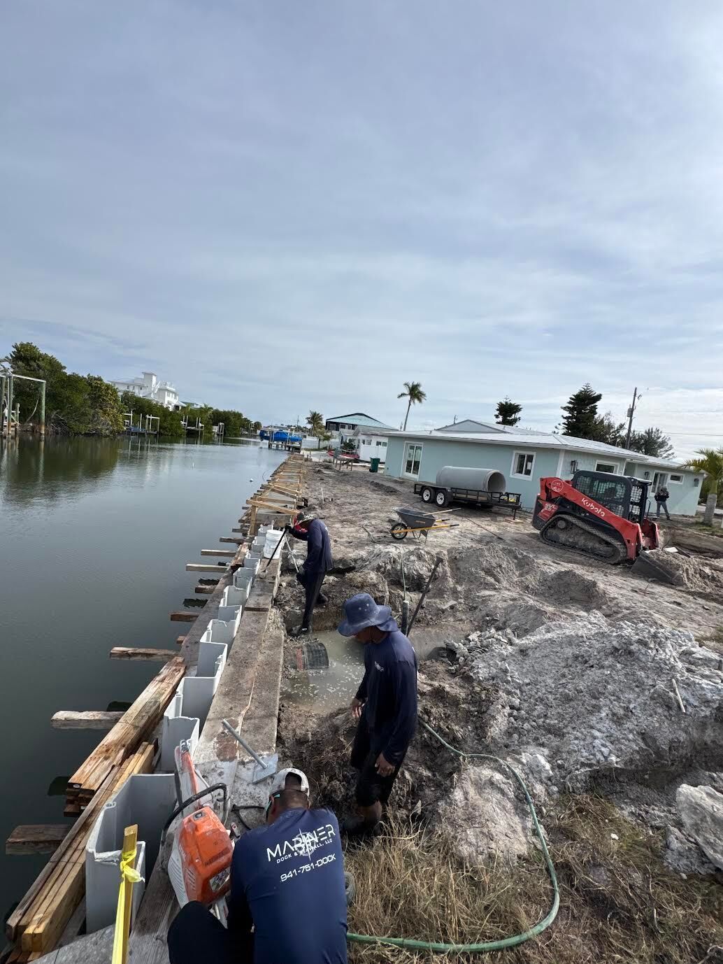 Workers repairing a riverbank with sandbags beside calm water and a small red tractor.