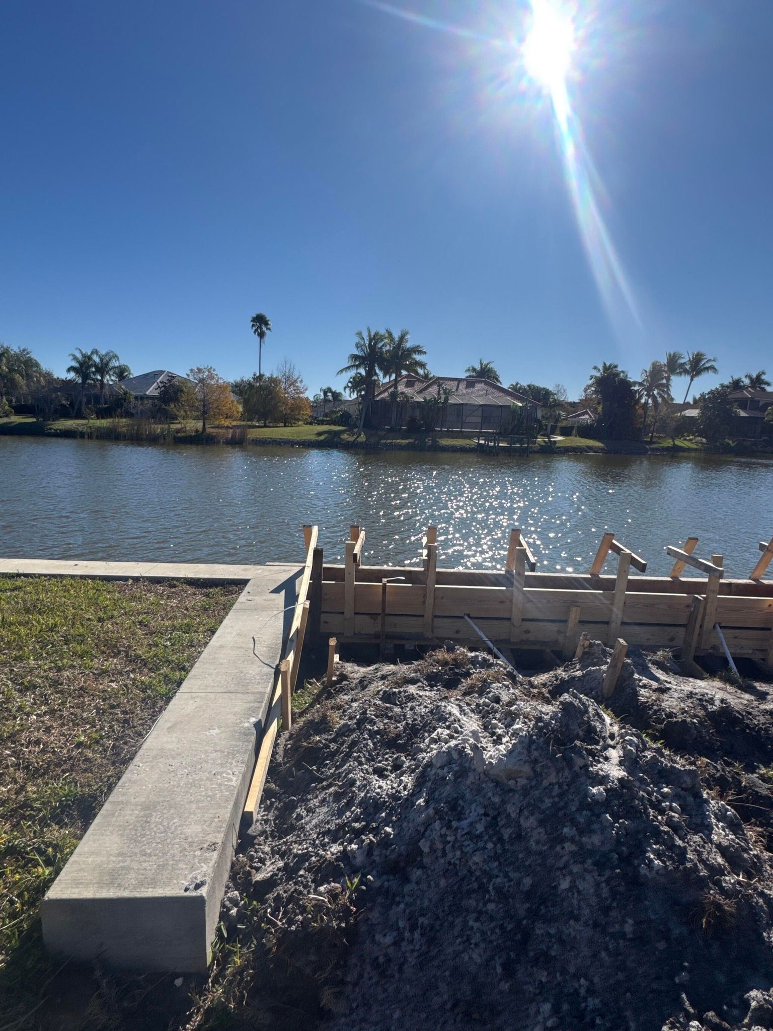 Sunny lakeside path beside a construction pile and wooden formwork, with palm trees across the water.