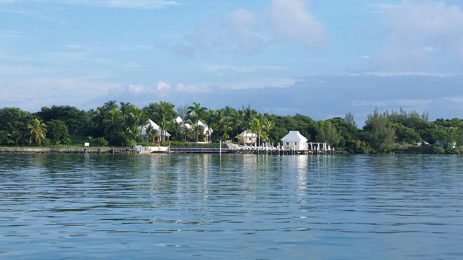 A row of white waterfront bungalows nestled among lush green trees along a calm, blue ocean shoreline.