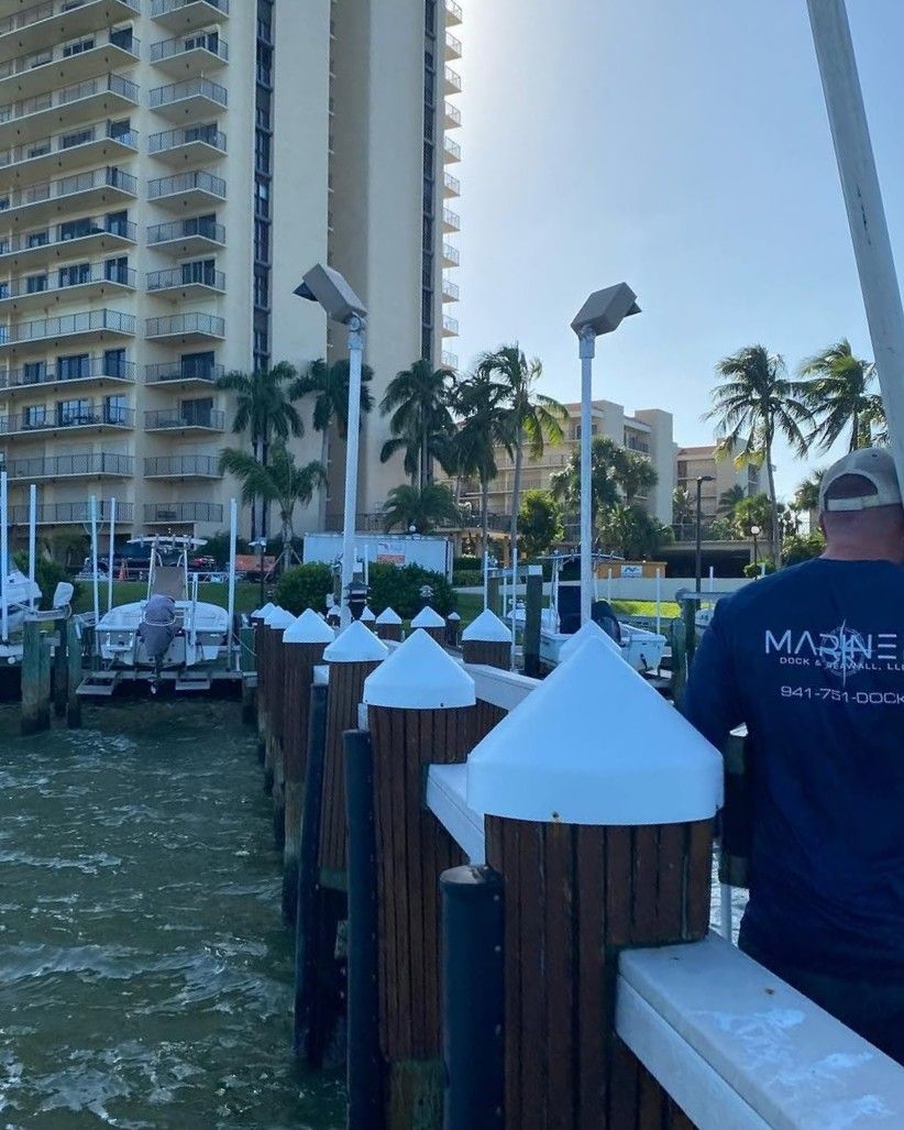 A person in a dark blue shirt stands on a dock by a marina, with tall residential buildings in the background.