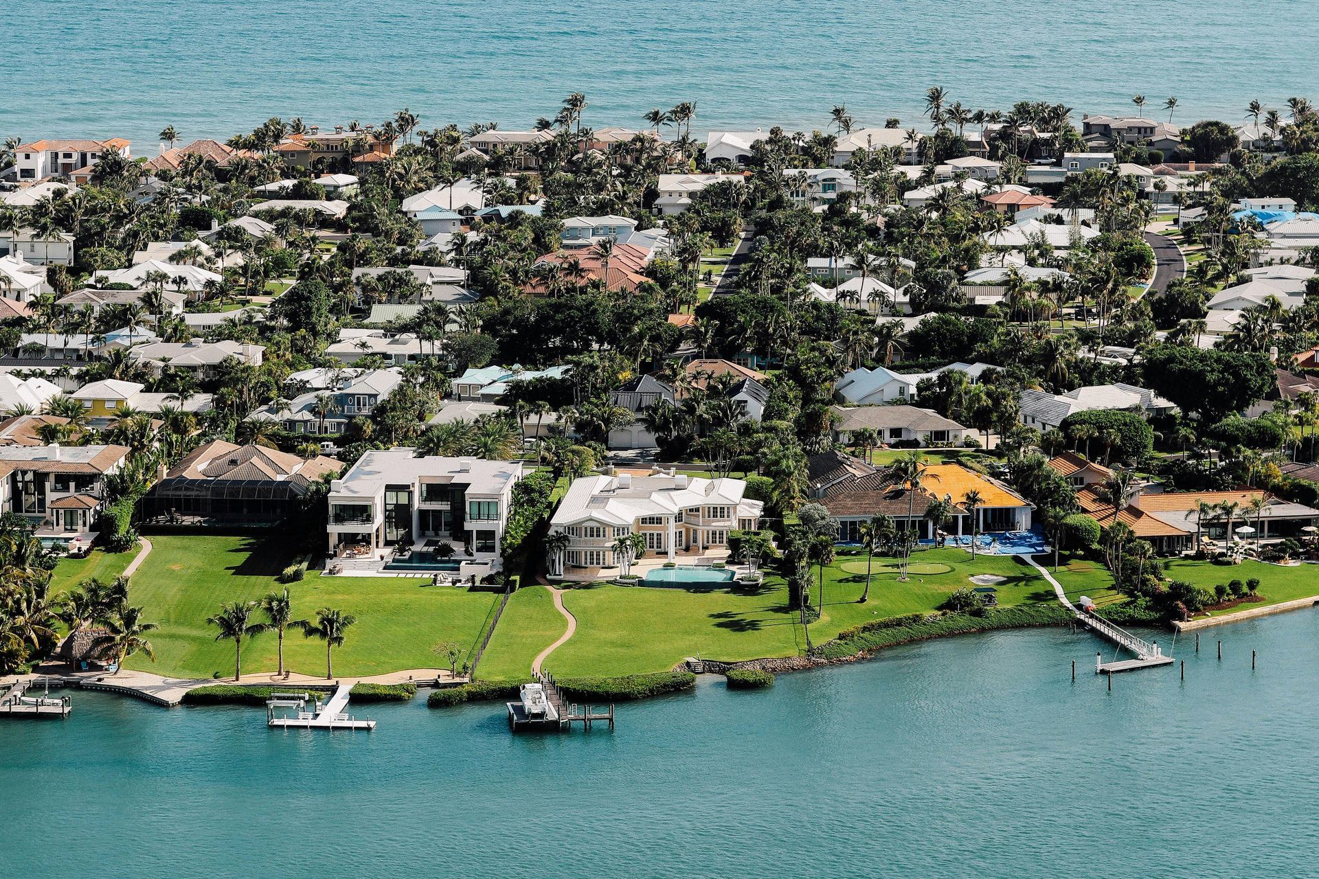 An aerial view of a coastal neighborhood with large luxury houses, green lawns, and private docks along a blue waterway.