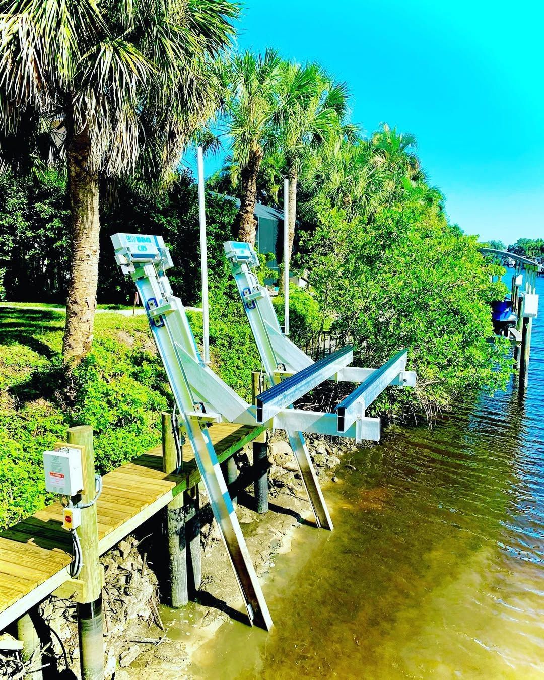 A metal boat lift system installed on a shoreline next to palm trees and a wooden walkway by the water.