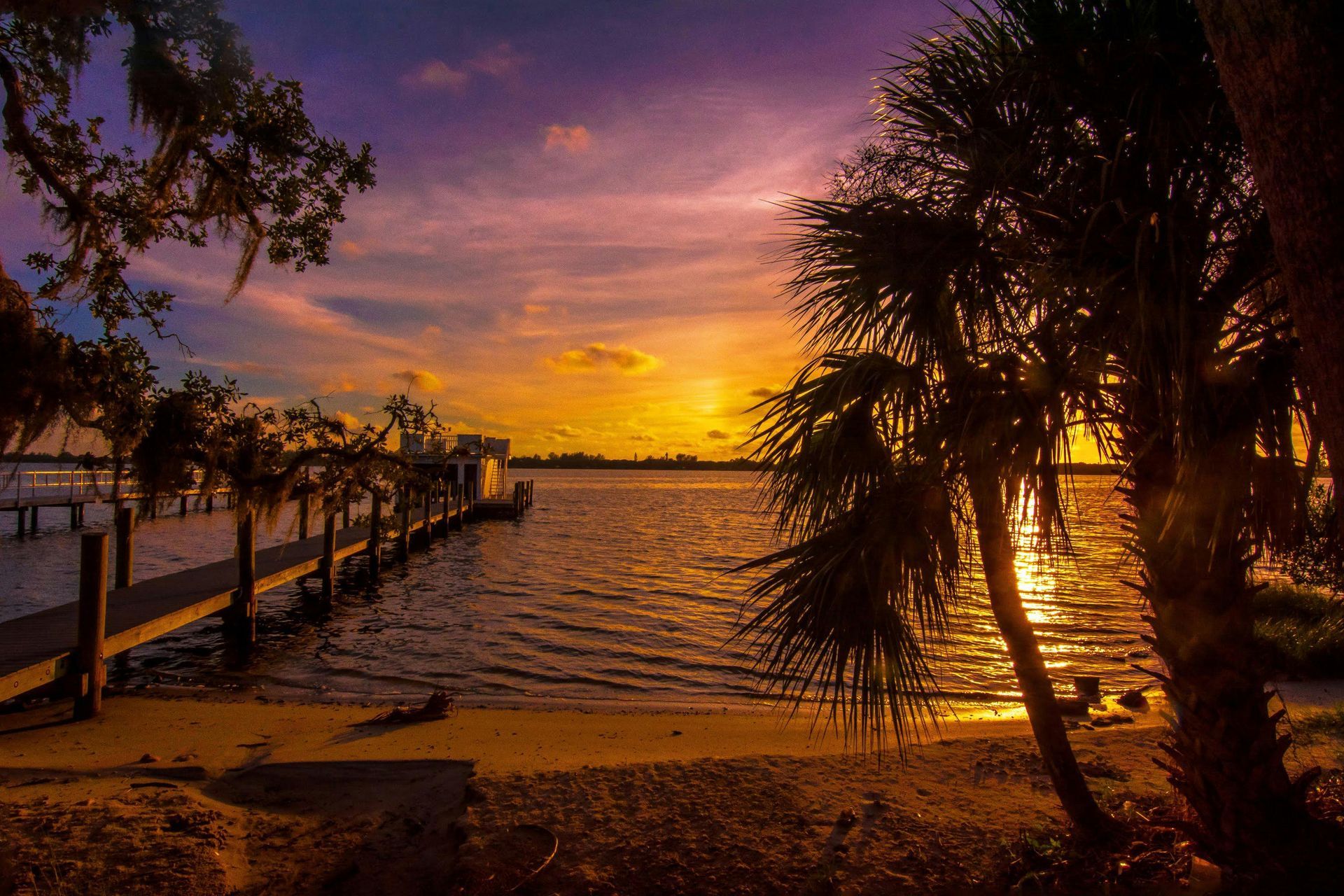 A pier extends into a calm lake under a vibrant sunset sky filled with purple, orange, and gold hues beside palm trees.