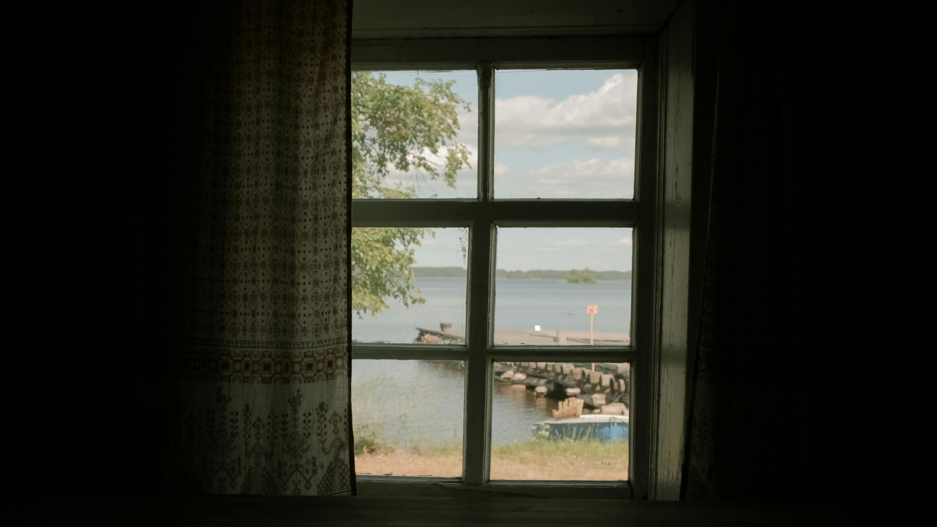 A view through a window showing a lake, a rocky shoreline, a small boat, and green trees under a bright blue sky.