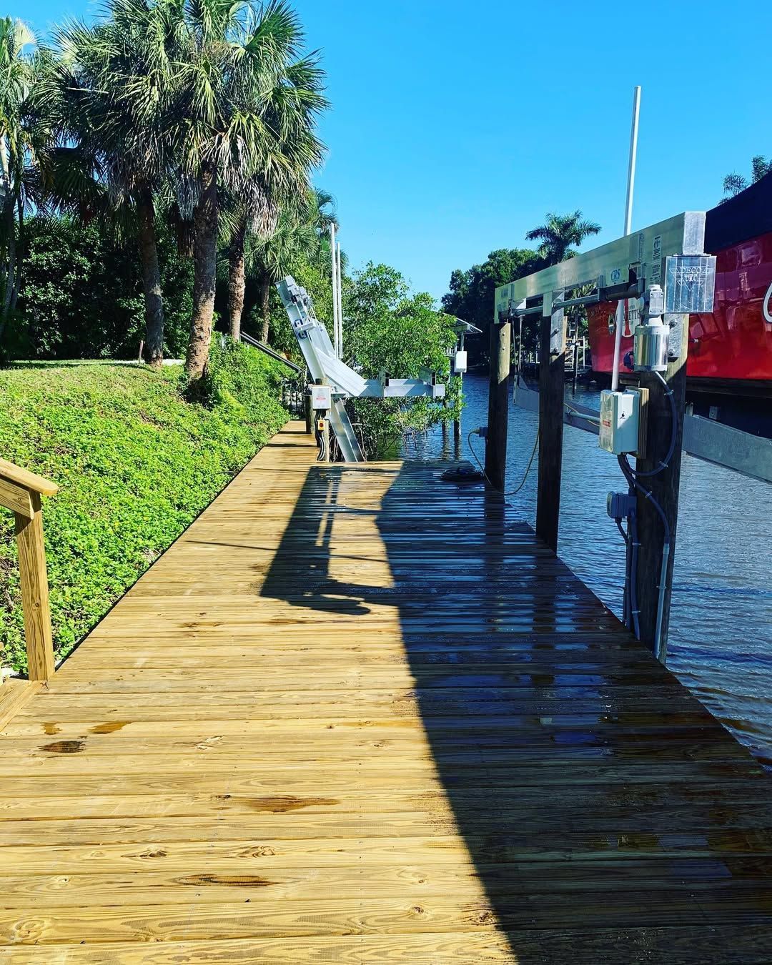 A wooden dock extends over calm water, framed by a sunlit grassy bank with palm trees and a boat lift structure.