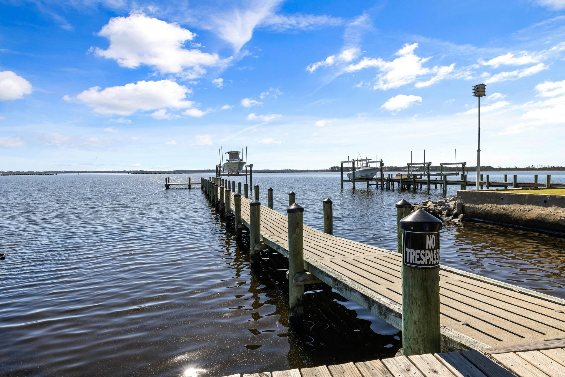 A wooden pier extends into a calm, blue waterway under a bright, sunny sky with clouds.