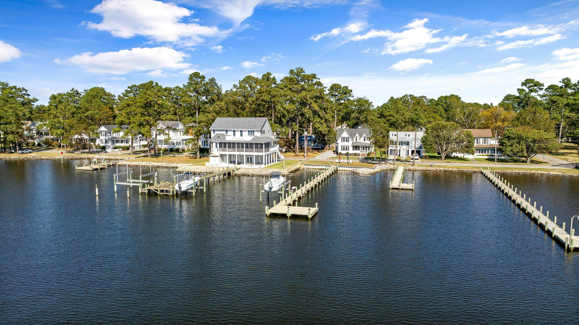 An aerial view of waterfront houses with wooden docks extending into a blue river under a bright, sunny sky.
