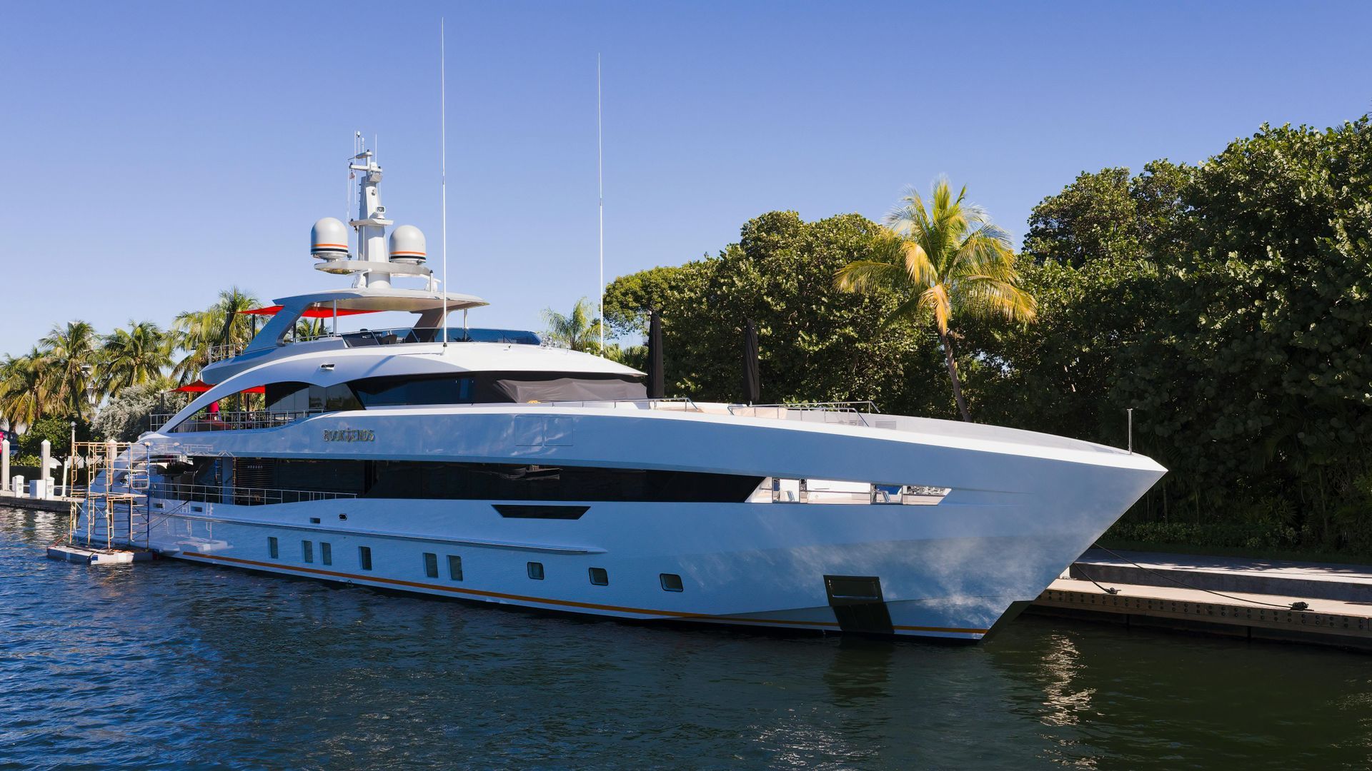 A sleek, modern white yacht is docked alongside a lush, green treeline under a clear blue sky.