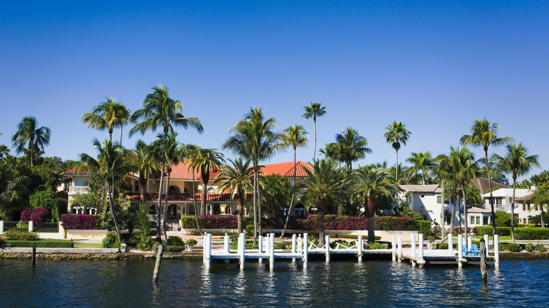 Waterfront homes with tile roofs and palm trees set against a clear blue sky, featuring a white wooden dock in the water.