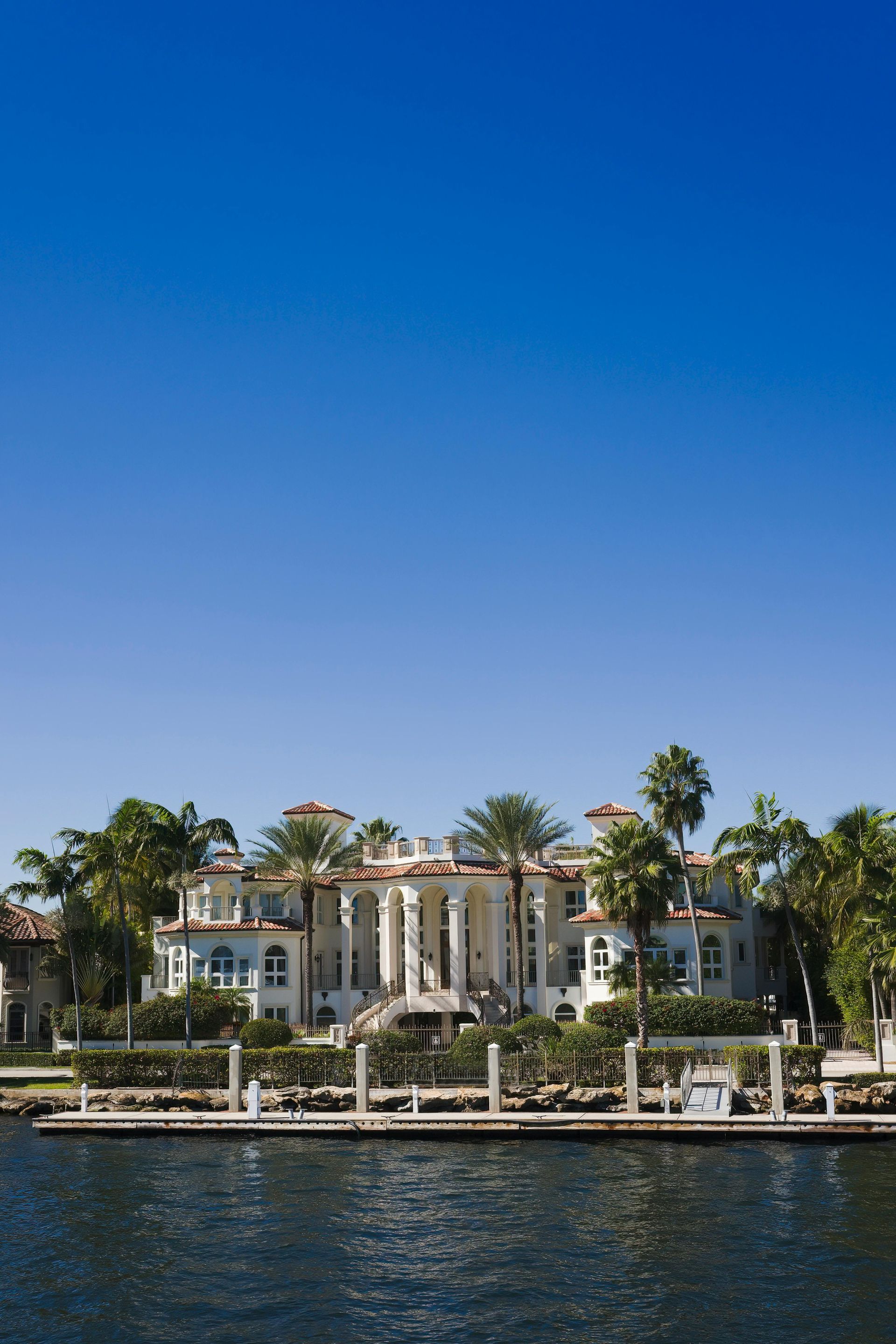 A large, white, multi-story mansion with classical columns and palm trees, situated on a waterfront under a clear blue sky.