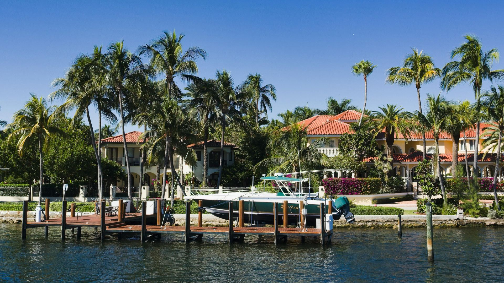 A boat docked at a wooden pier in front of tropical waterfront houses under a clear blue sky.