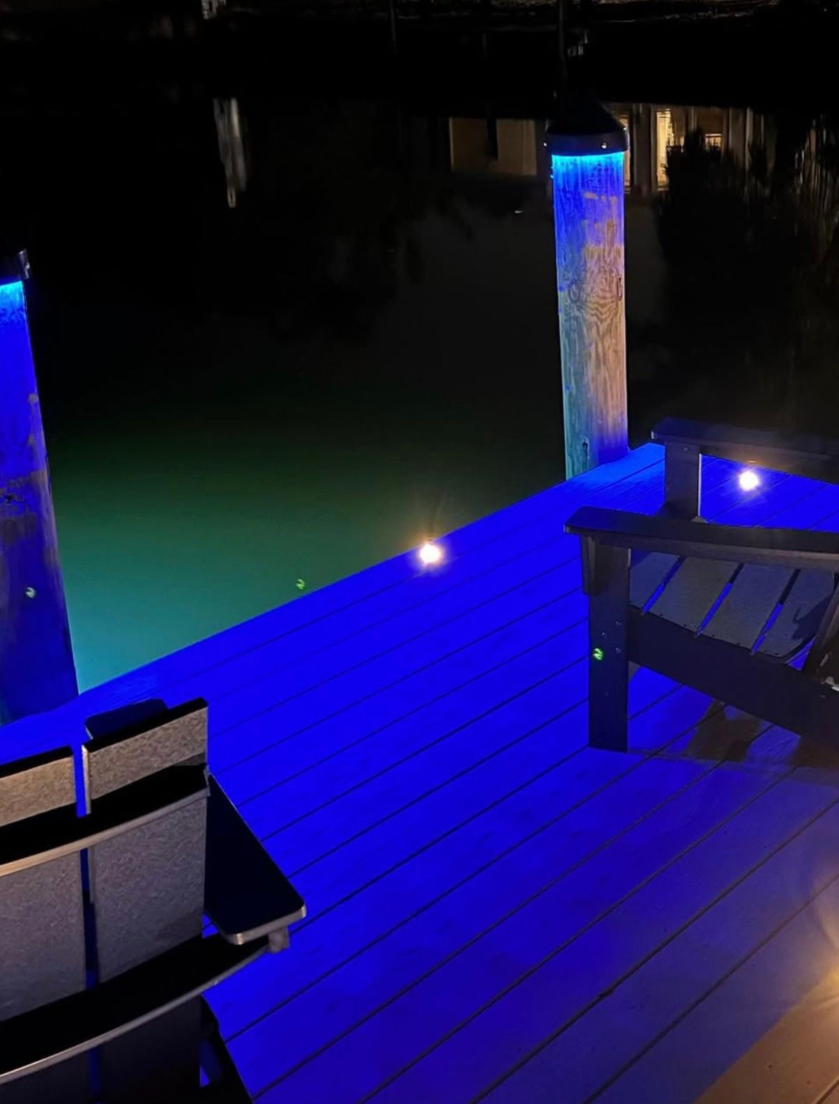 A wooden dock at night with blue deck lighting, two dark chairs, and illuminated pilings overlooking dark water.