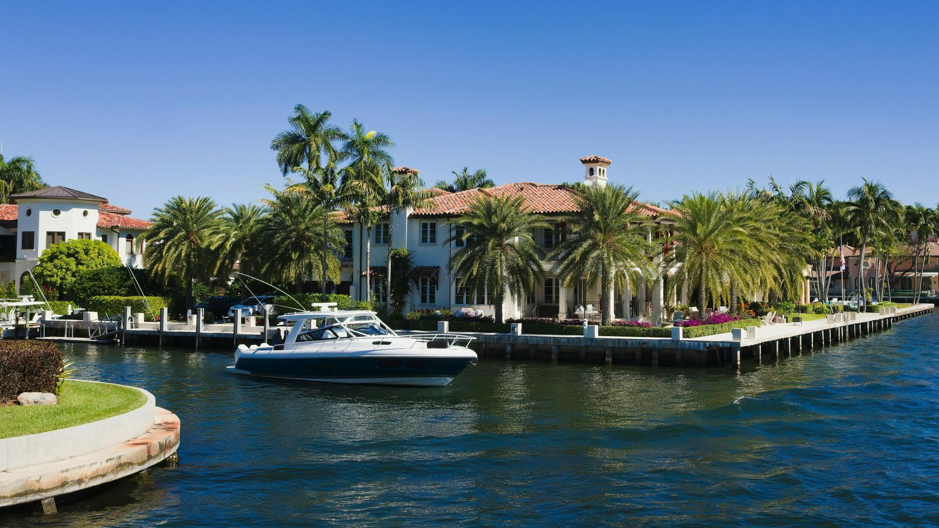 A motorboat travels through a sunny canal lined with large Mediterranean-style mansions and palm trees.