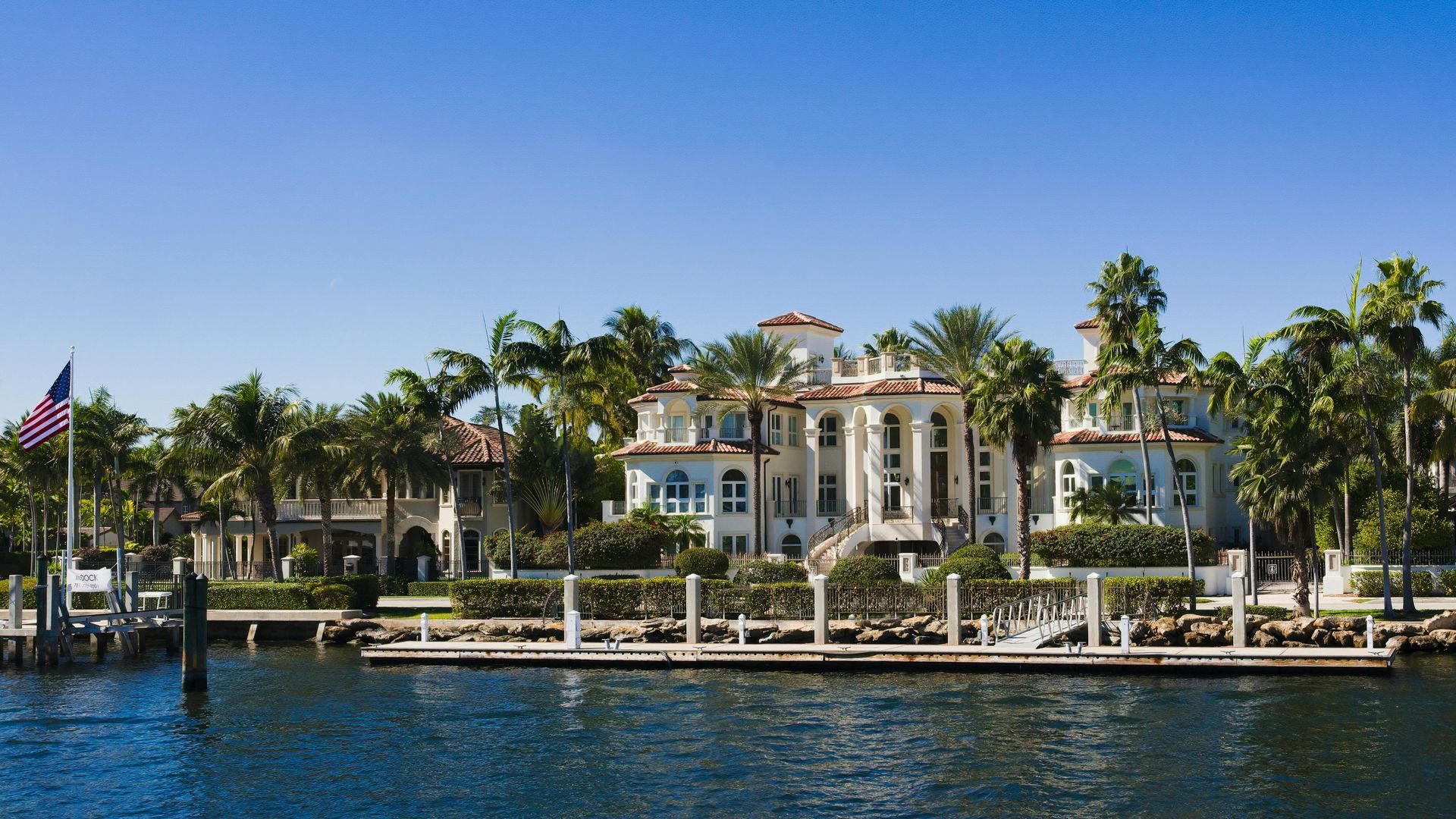 A large, multi-story white villa sits on a waterfront property, surrounded by palm trees with a wooden dock in the front.