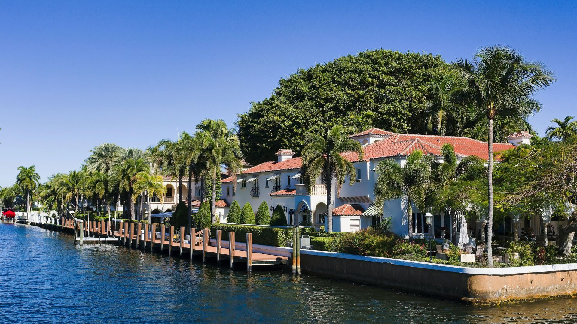 A white waterfront mansion with a red roof sits along a calm canal, lined by a wooden dock and numerous palm trees.