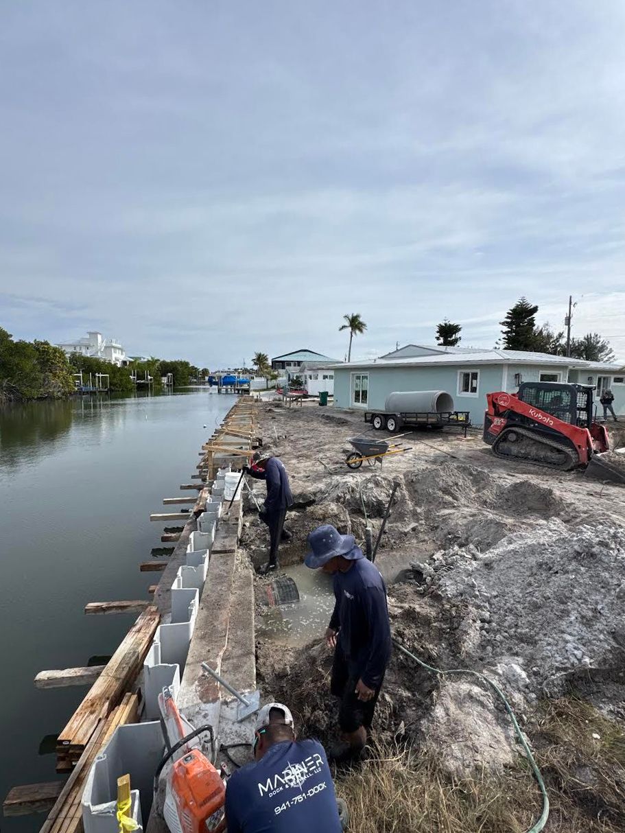 Construction workers install a concrete seawall along a canal, using tools near machinery under a bright, overcast sky.