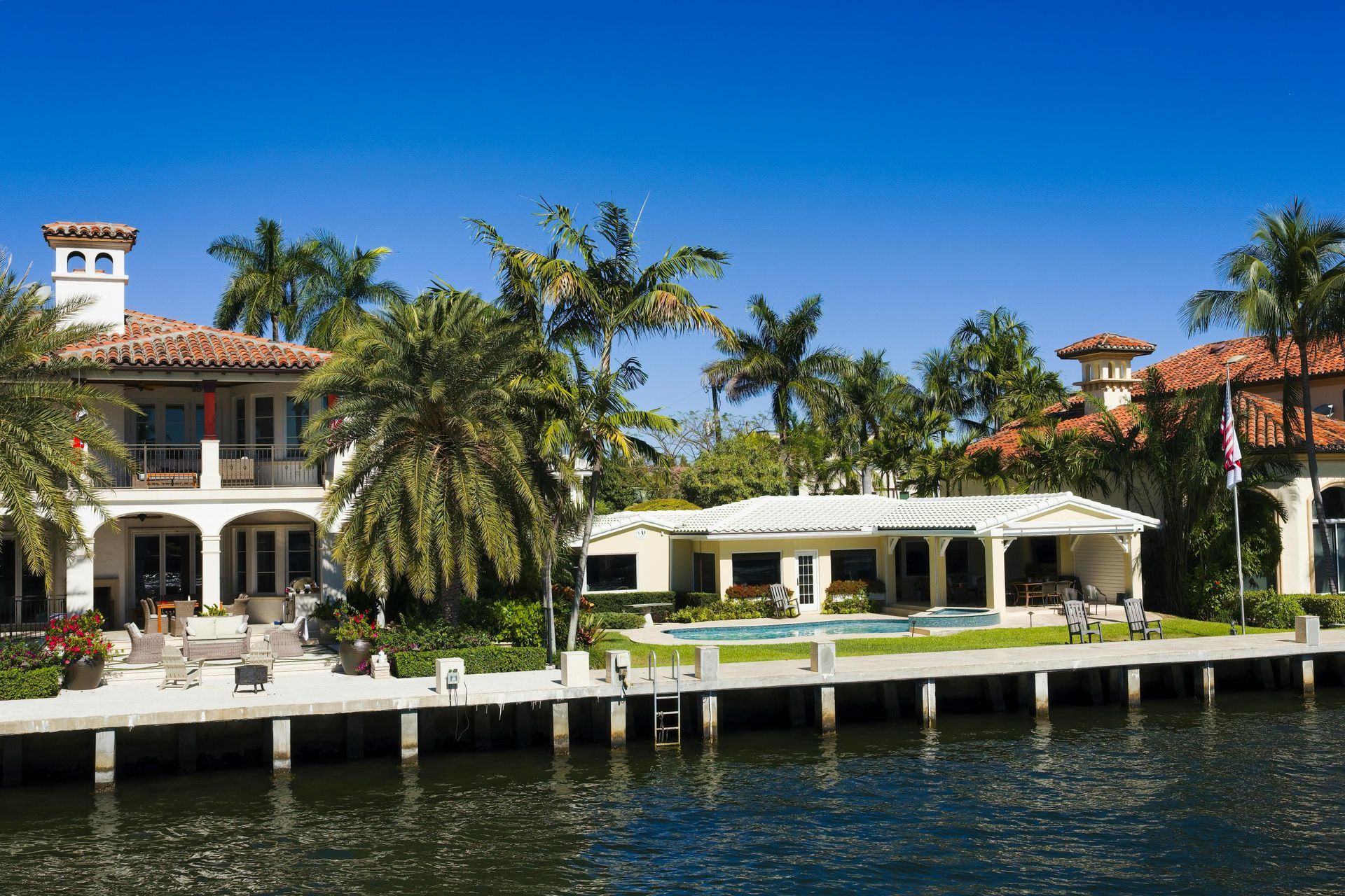 A luxurious waterfront home with a swimming pool, palm trees, and a wooden dock under a clear blue sky.