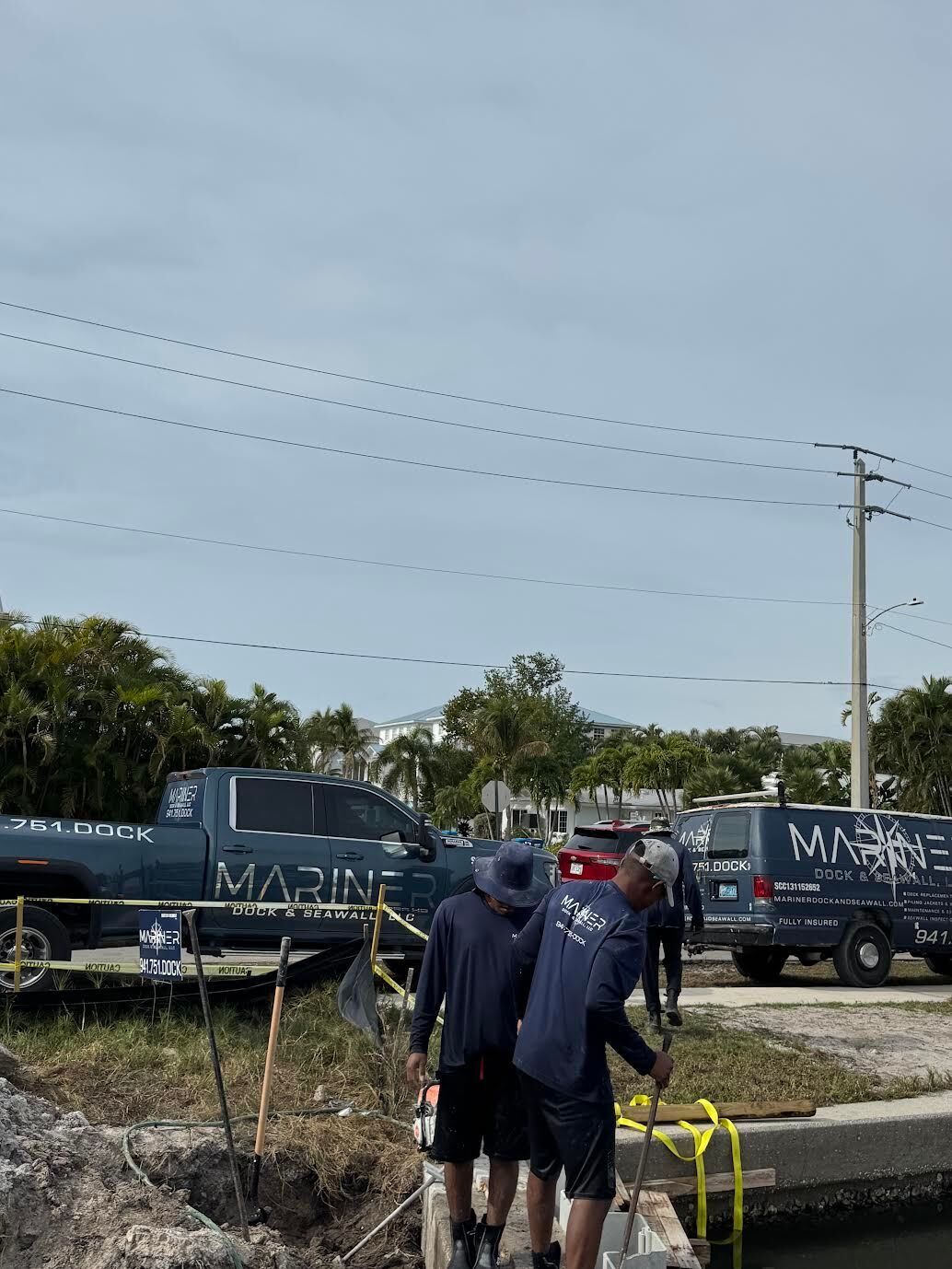 Two workers in dark uniforms stand by a canal next to two trucks marked with 