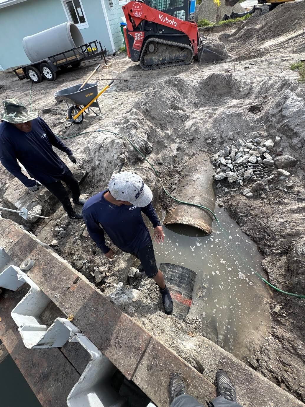 Two workers in a construction pit with exposed pipes, a red skid steer, and a trailer with a large pipe nearby.