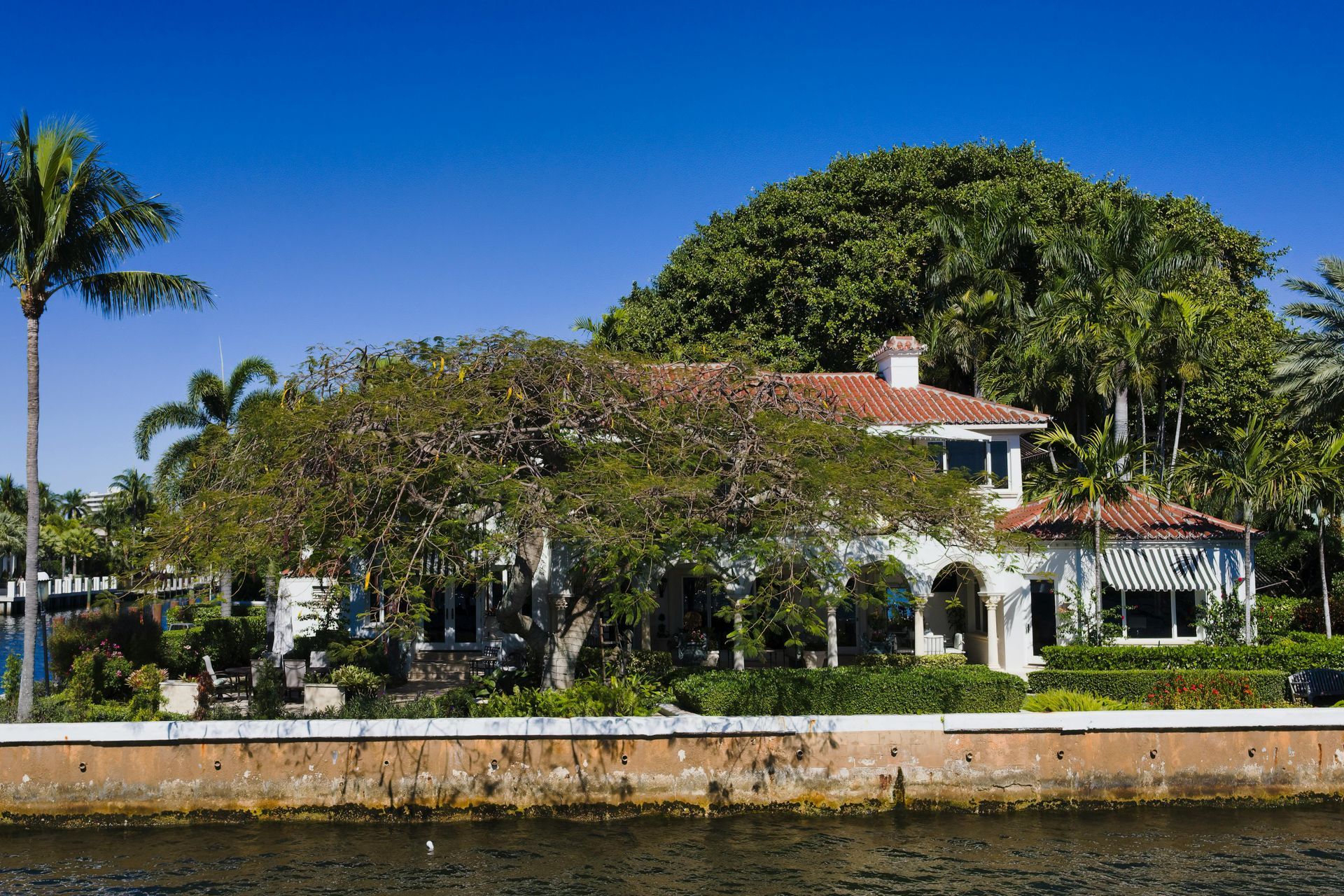 A white house with a red tile roof surrounded by lush trees and palms, situated behind a seawall along a canal.