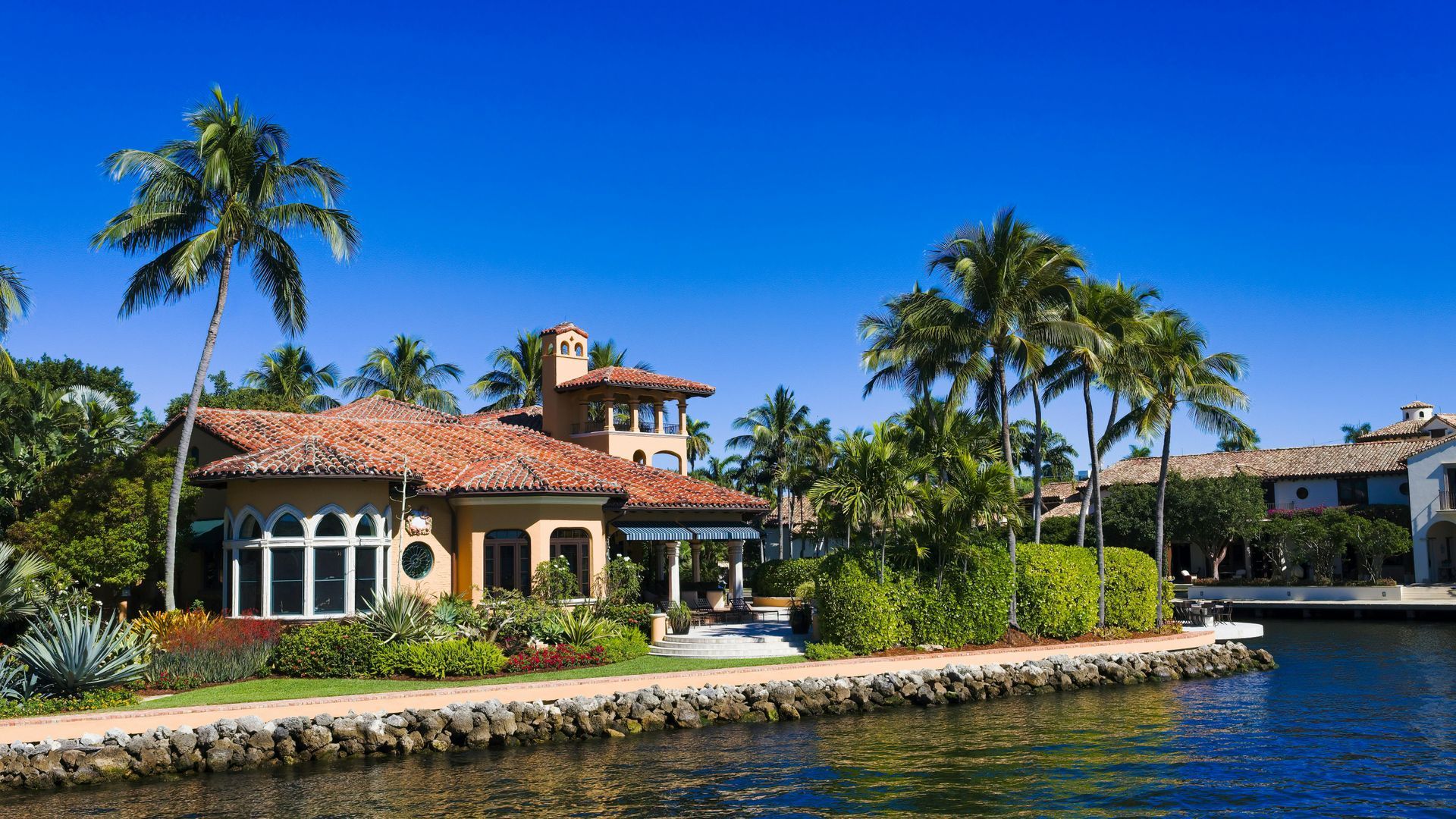 A tan house with a terracotta roof, surrounded by palm trees and lush landscaping along a calm, sunny canal.