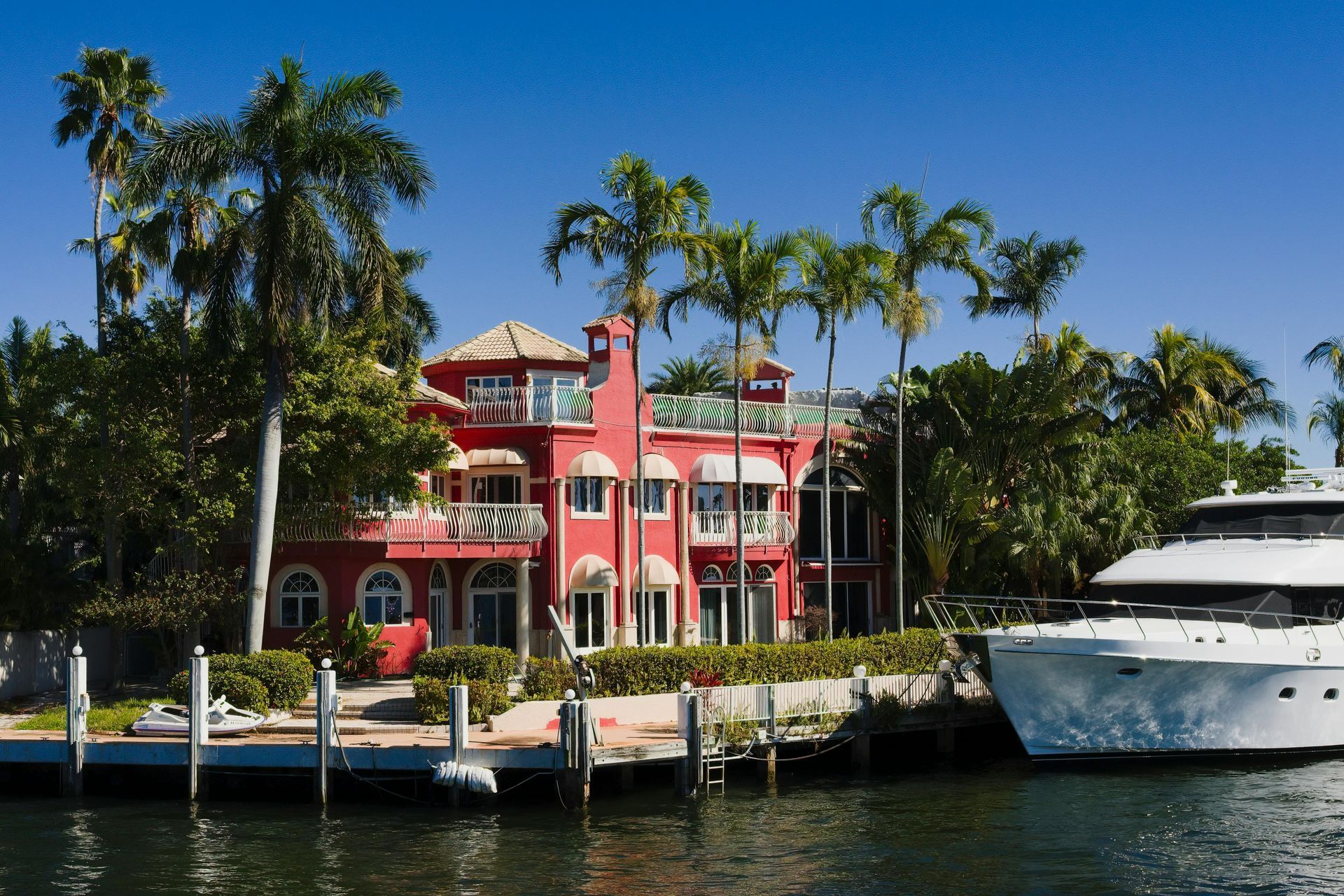 A vibrant red waterfront mansion surrounded by palm trees with a large white yacht docked in the foreground.
