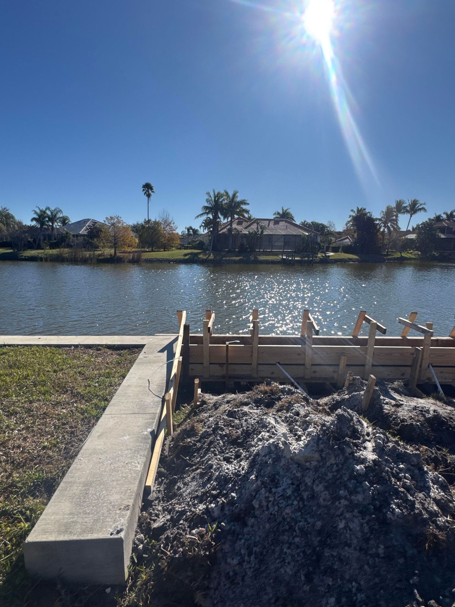 Construction forms and a pile of dirt sit near a concrete edge by a sunny waterfront with homes in the background.