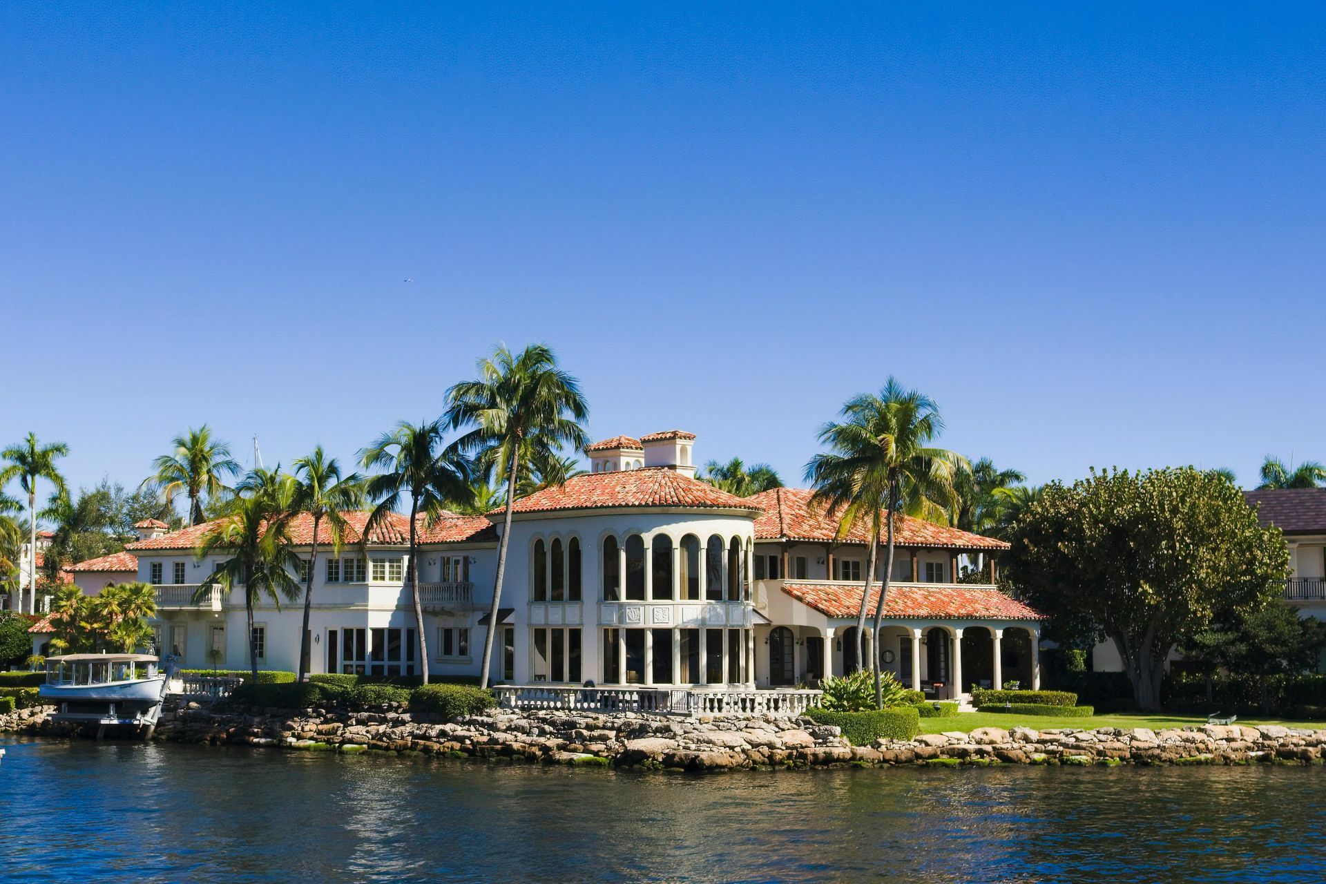 A two-story, white Mediterranean-style waterfront villa with a red-tiled roof, surrounded by palm trees under a blue sky.