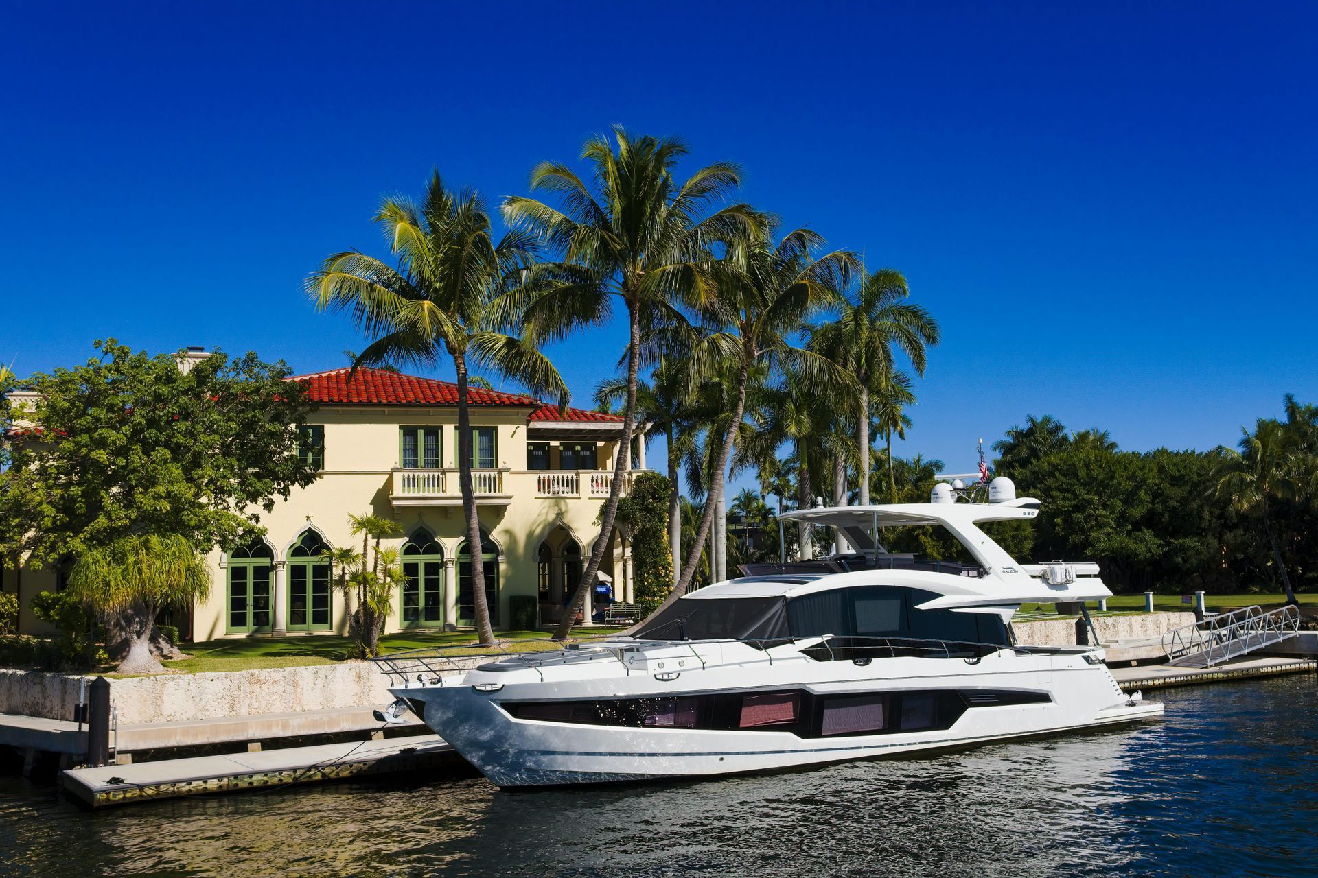 A modern white yacht docked at a private residence with a red-tiled roof and tall palm trees against a clear blue sky.