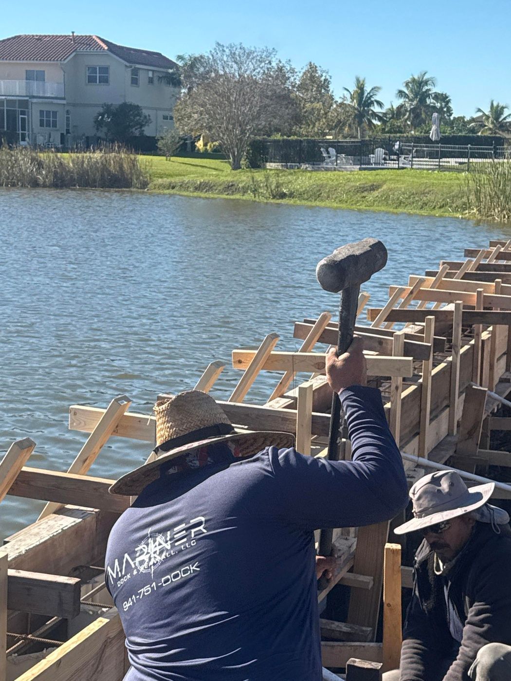 Two workers in sun hats and long sleeves build a wooden retaining wall along a waterfront with a sledgehammer.