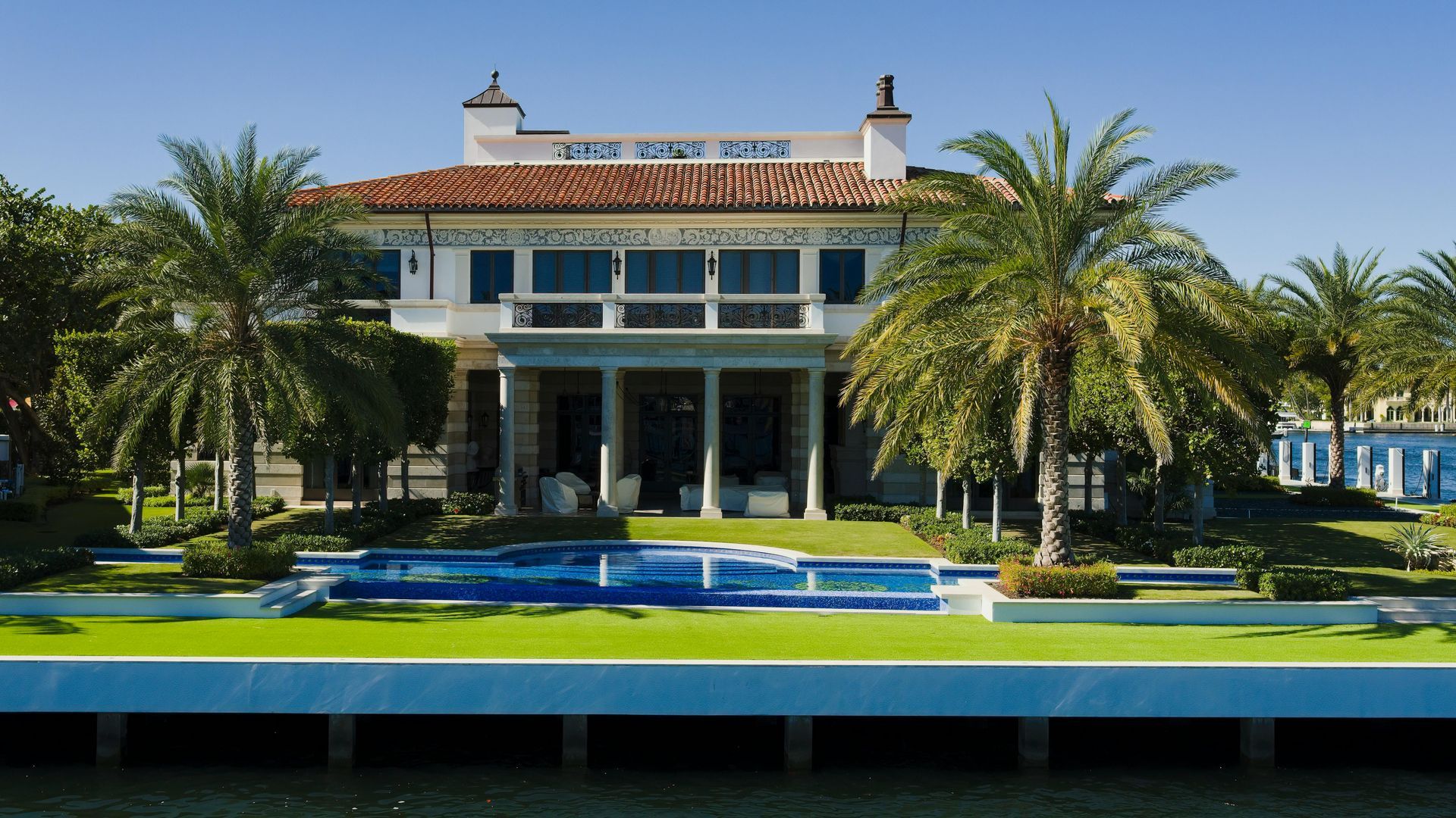 A Mediterranean-style waterfront mansion with a tiled roof, palm trees, and a pool, viewed from across a canal.