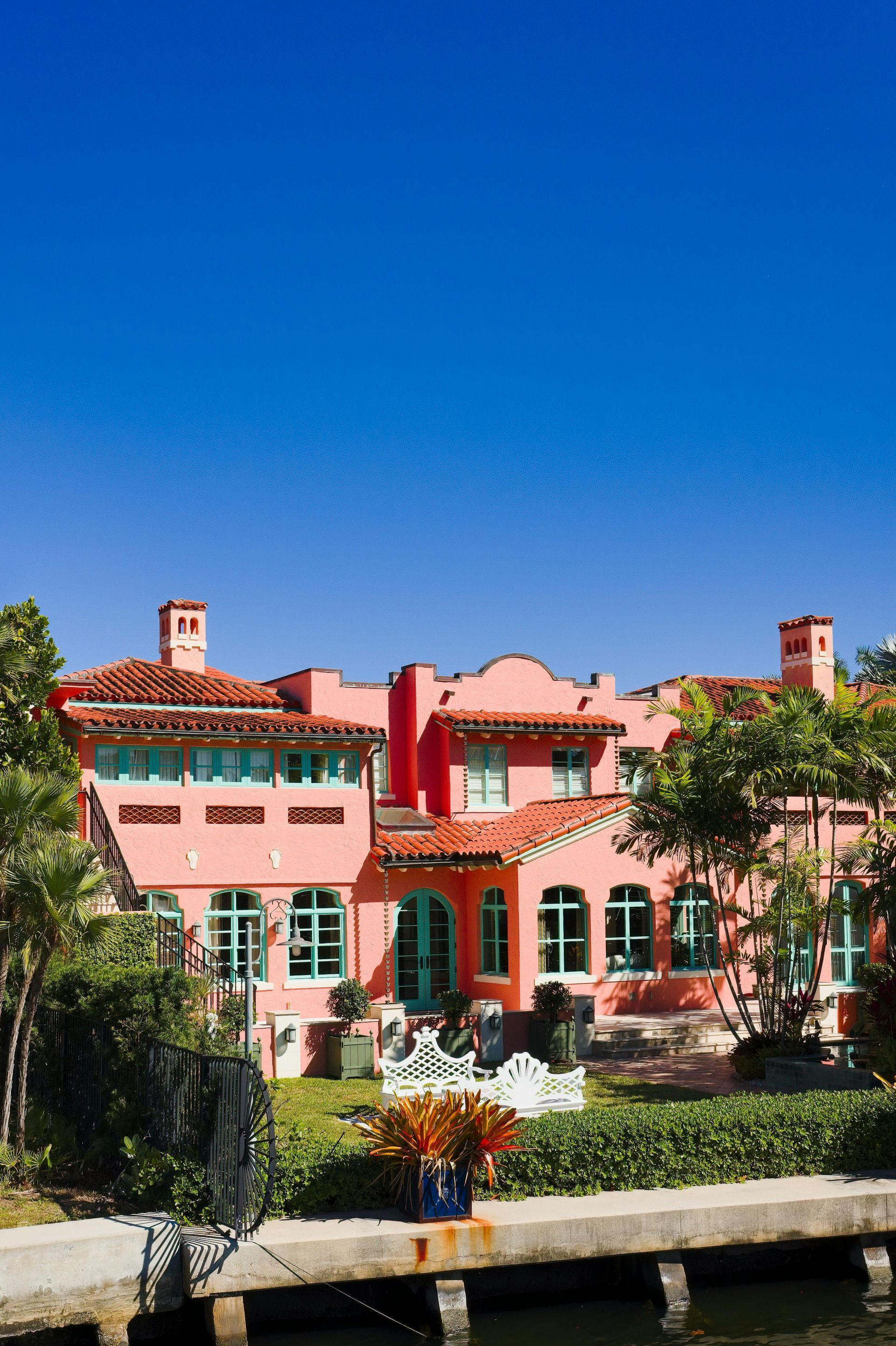A pink Spanish-style villa with a terracotta roof, set against a bright blue sky behind a garden and a waterfront wall.