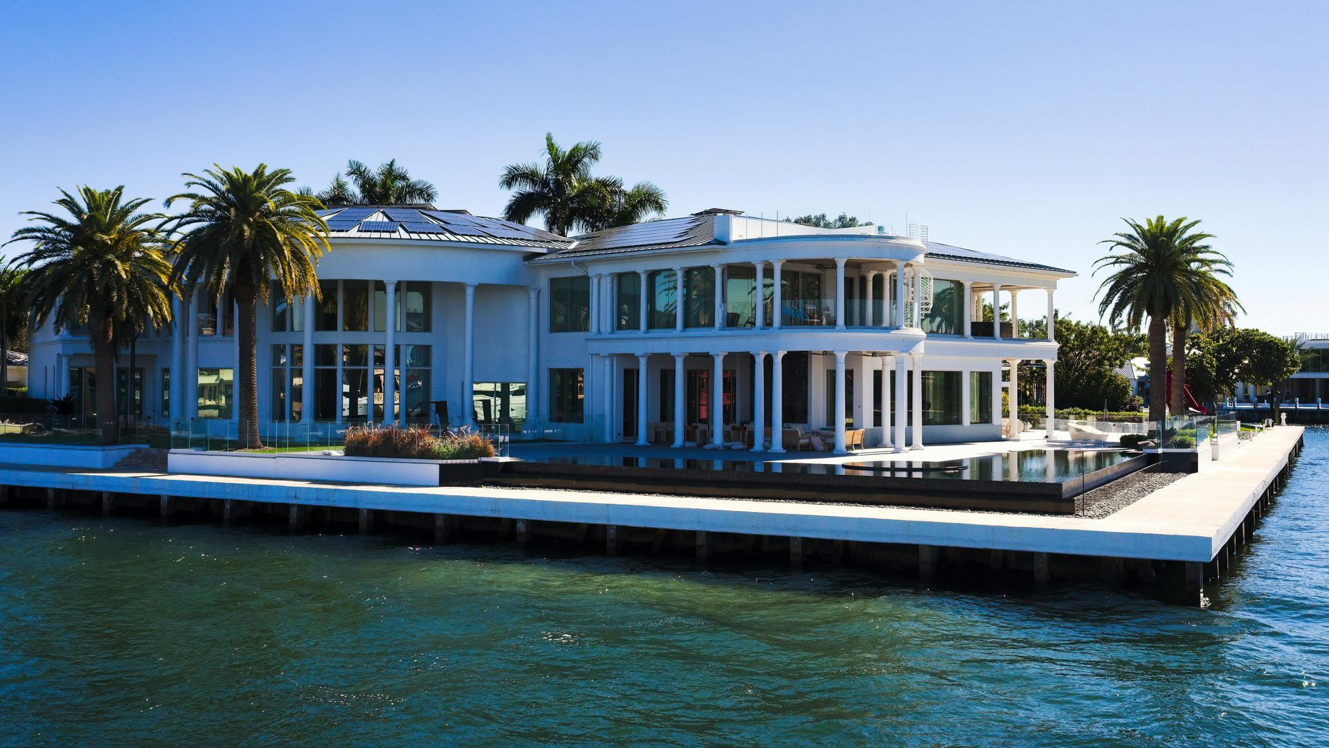 A large, two-story white modern house with tall columns and palm trees, situated directly on the water.