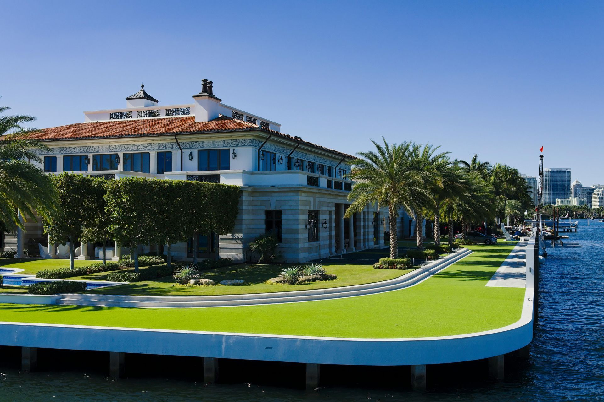 A white, mediterranean-style building with a terracotta roof sits on a waterfront lot with manicured green lawns and palms.