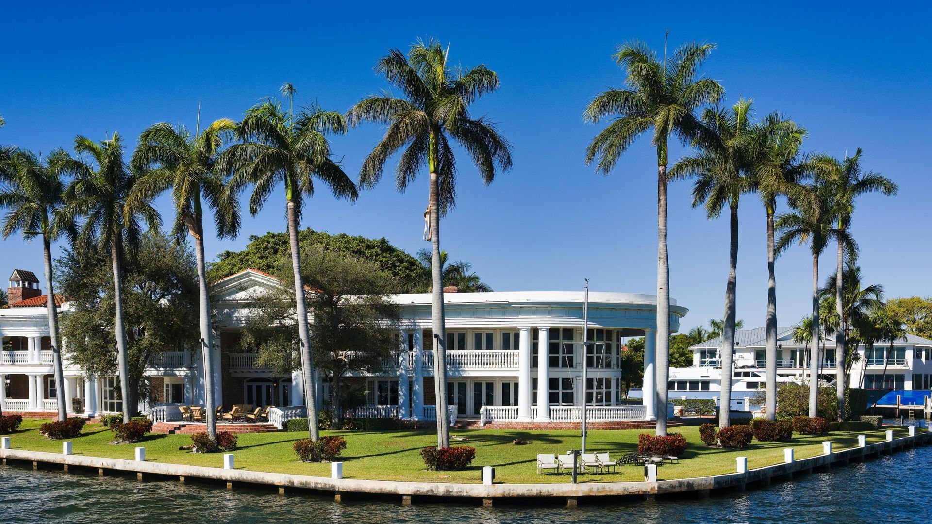 A white, multi-story building with tall columns stands on a grassy waterfront, surrounded by several tall palm trees.