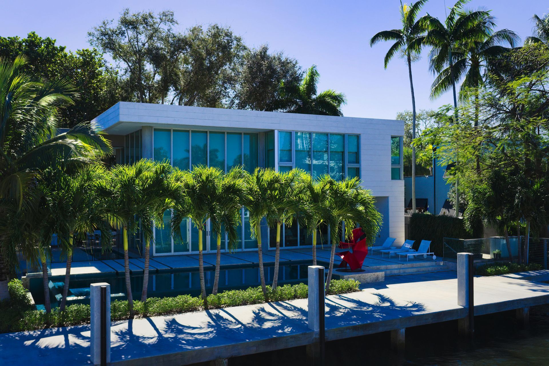 A two-story modern house with large windows, a swimming pool, and a row of palm trees next to a boat dock.