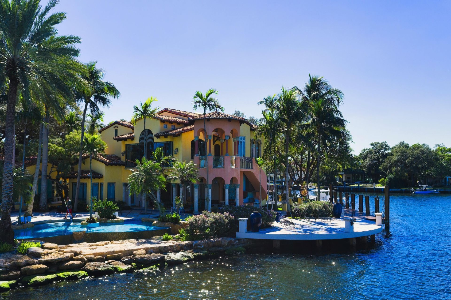 A Mediterranean-style house with yellow and peach walls, a swimming pool, and palm trees beside a blue canal.