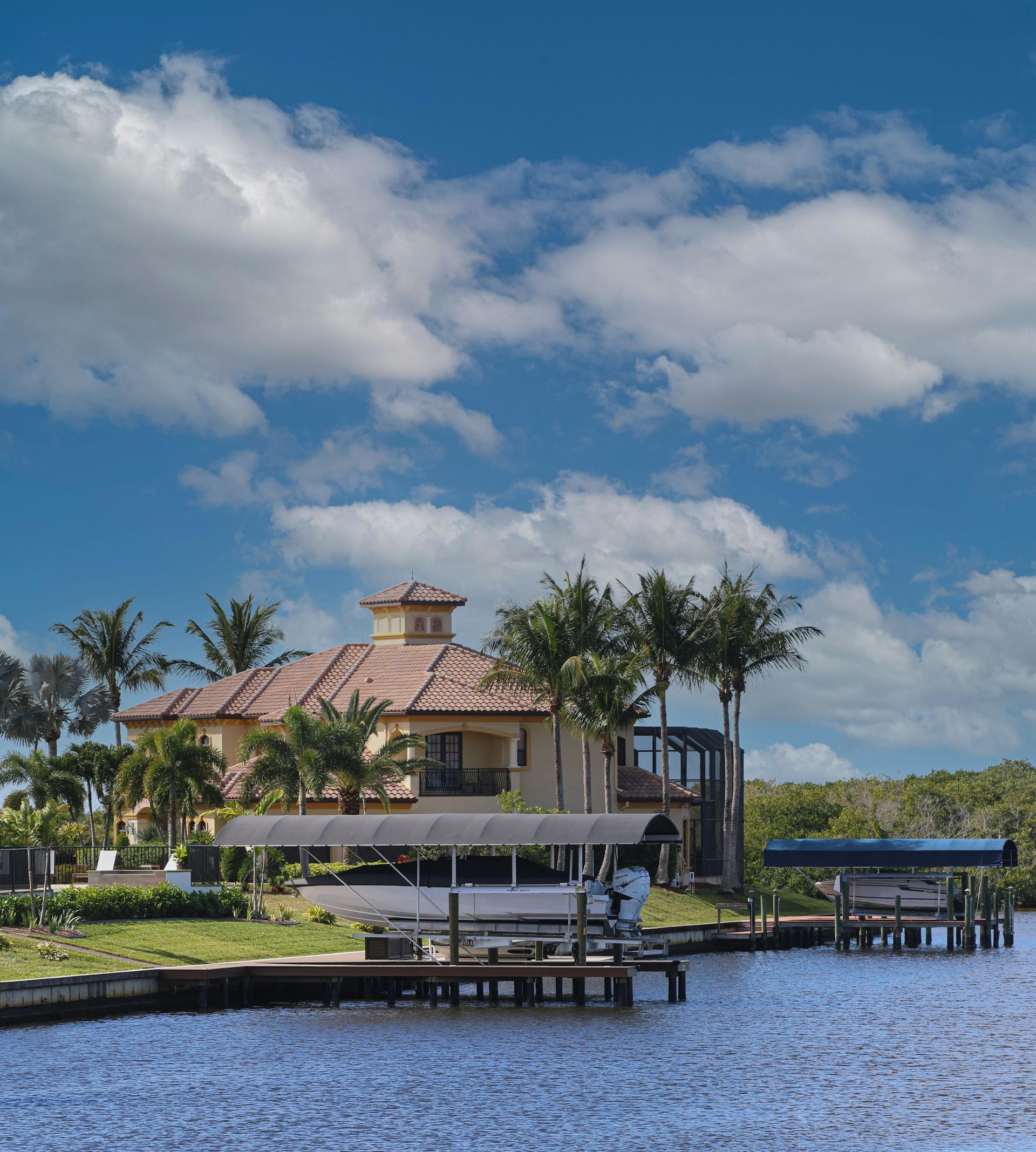 A tan mansion with a tiled roof sits on a waterfront lot with palm trees, boat docks, and blue skies with clouds.
