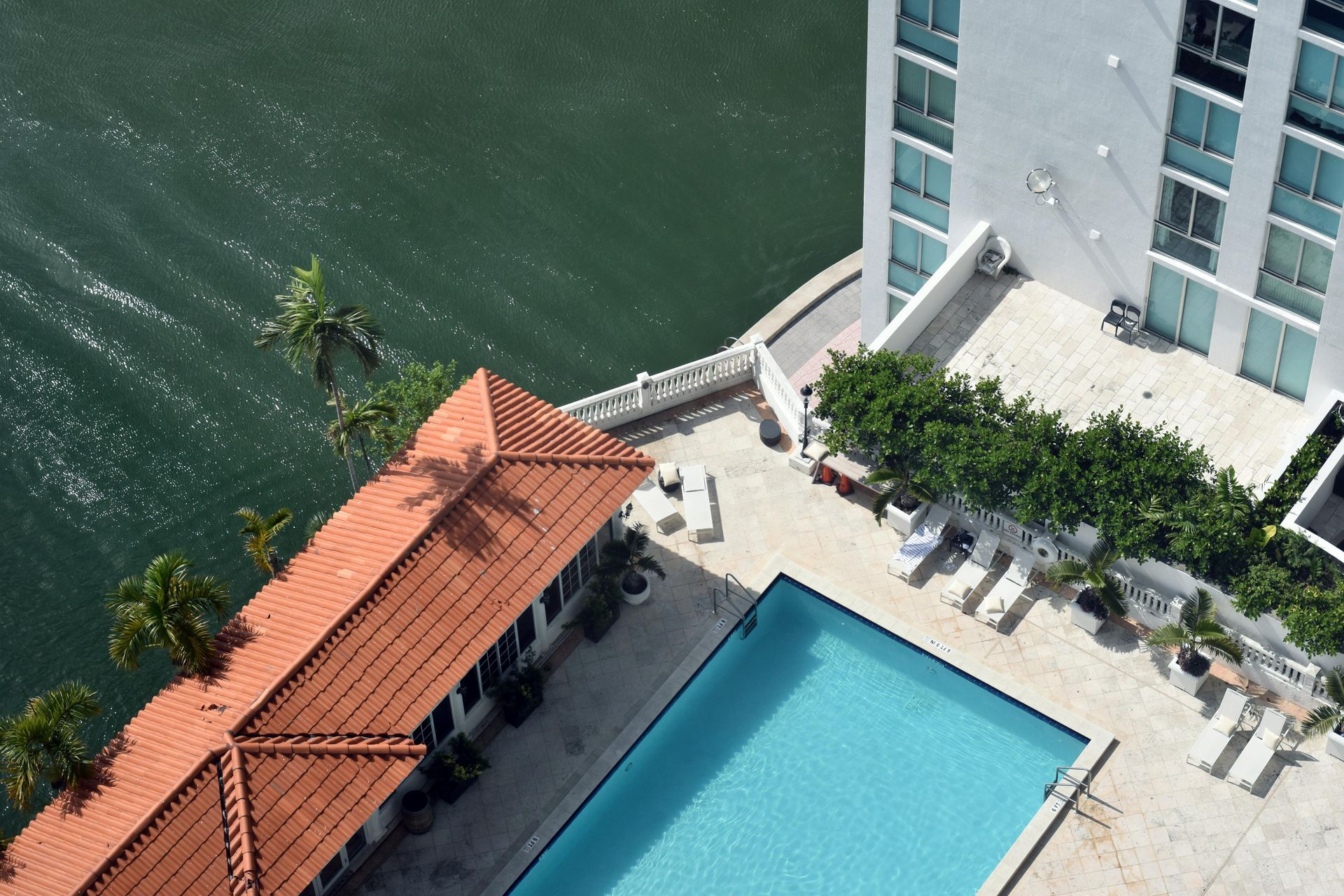 An aerial view of a rectangular swimming pool next to a building with a terracotta-tiled roof and a body of water.