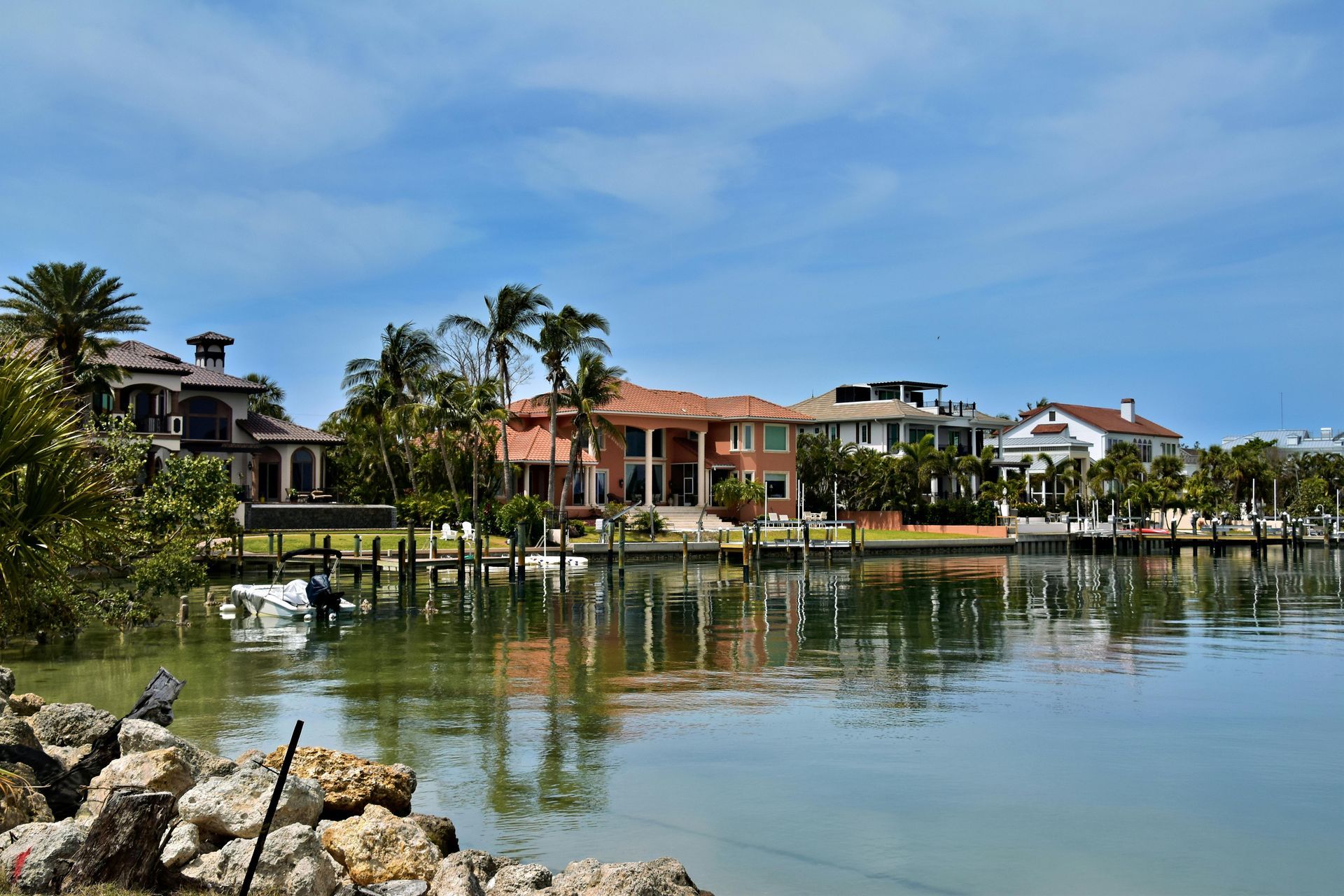 Large luxury waterfront homes along a calm canal with palm trees and a rocky shoreline in the foreground under a blue sky.
