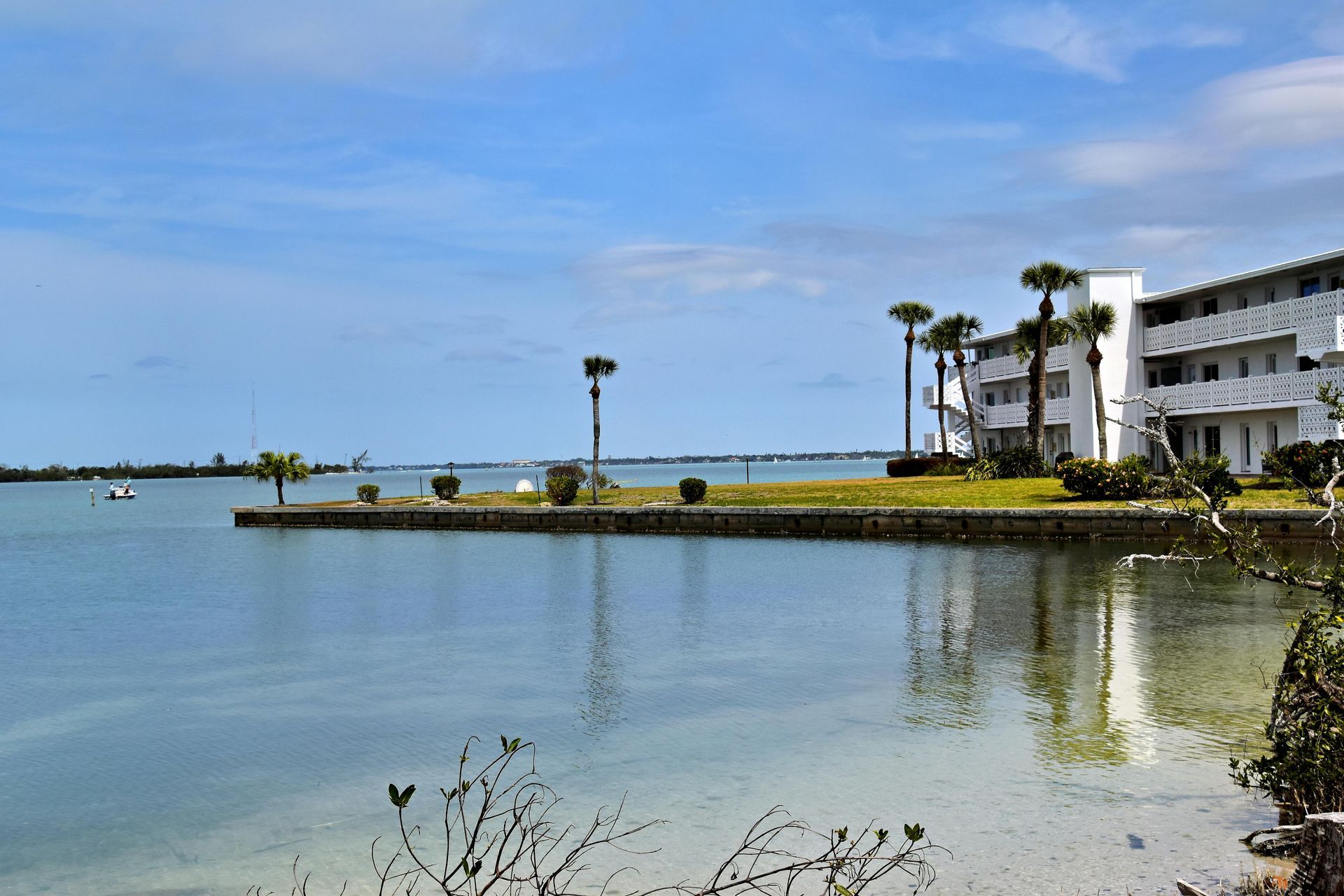 A white multi-story apartment building sits on a grassy shore along a calm, clear bay under a blue sky.